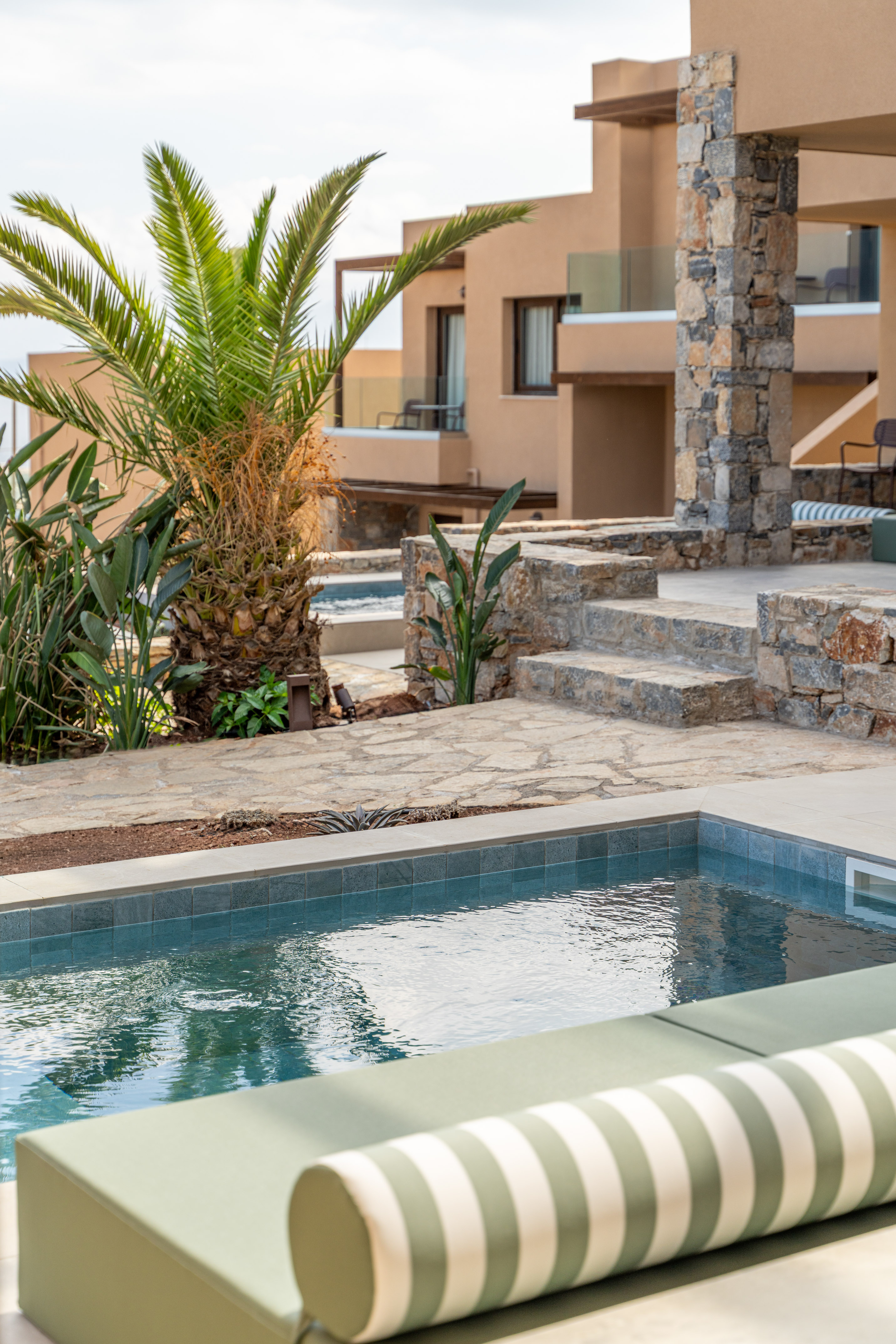 Private pool, green striped sunbed, palm tree, and modern resort building.