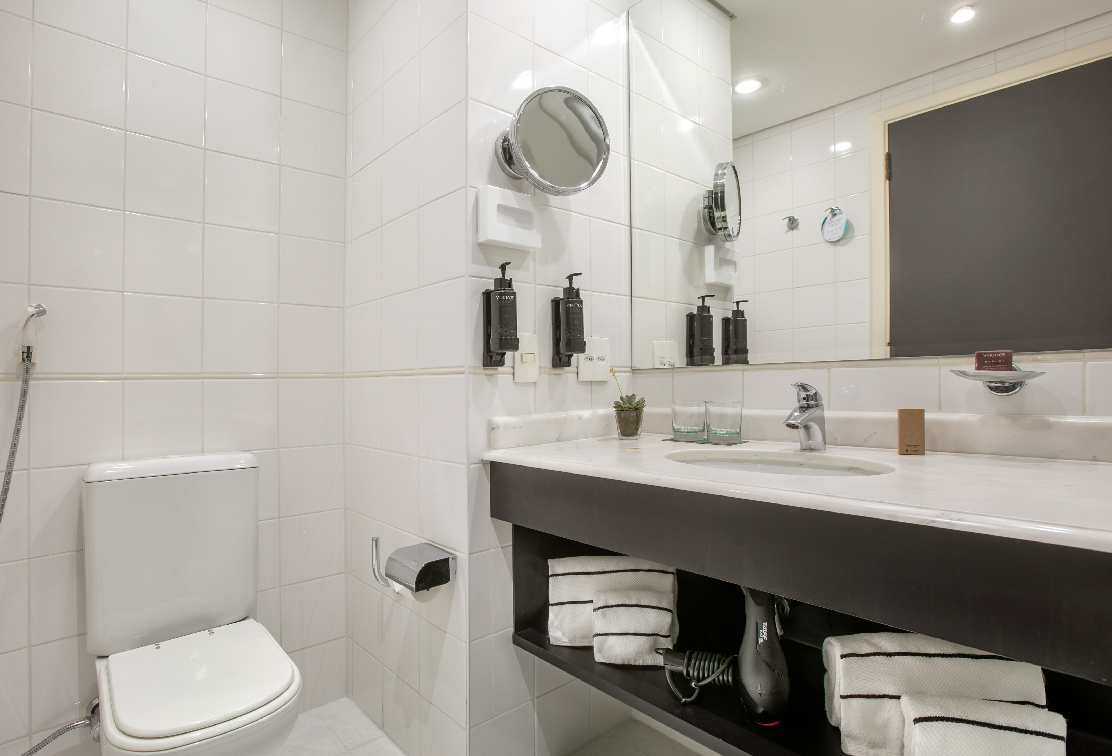 Modern hotel bathroom with white tiled walls, vanity, sink, mirror, and toilet.