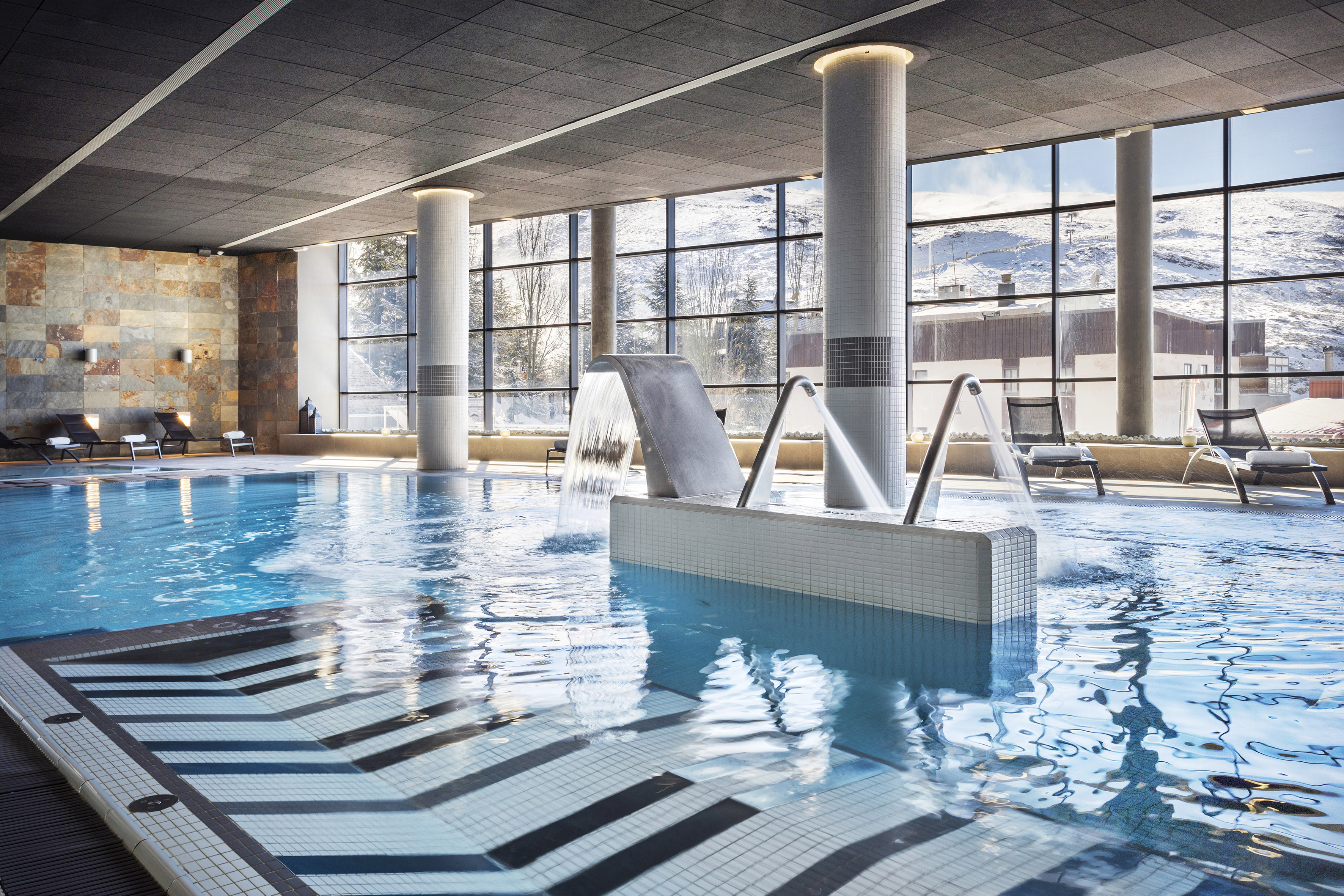 Indoor spa pool with waterfall, jets, and large windows overlooking snowy mountains.