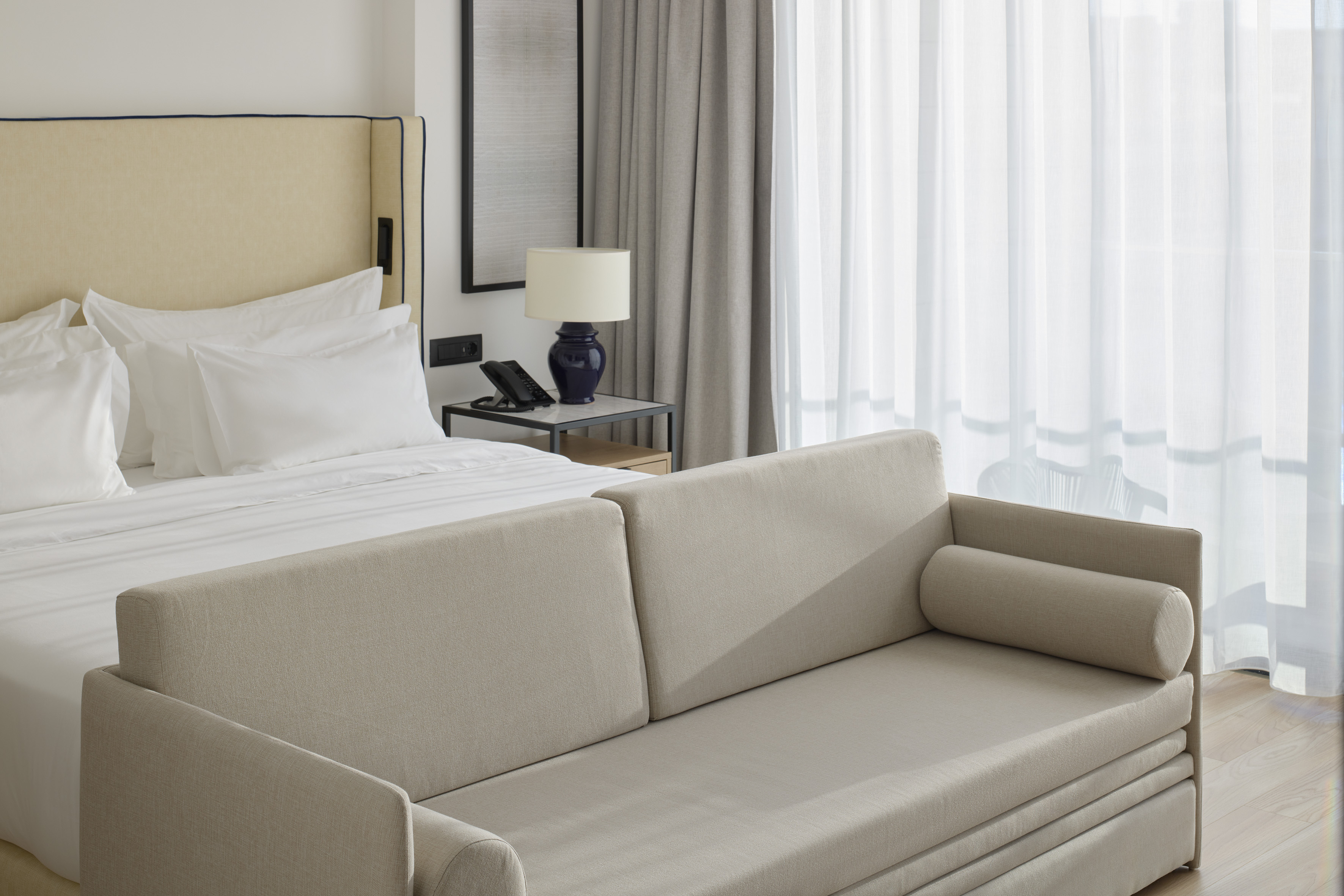 Modern hotel room with bed, beige sofa, bedside lamp, and sheer curtains.