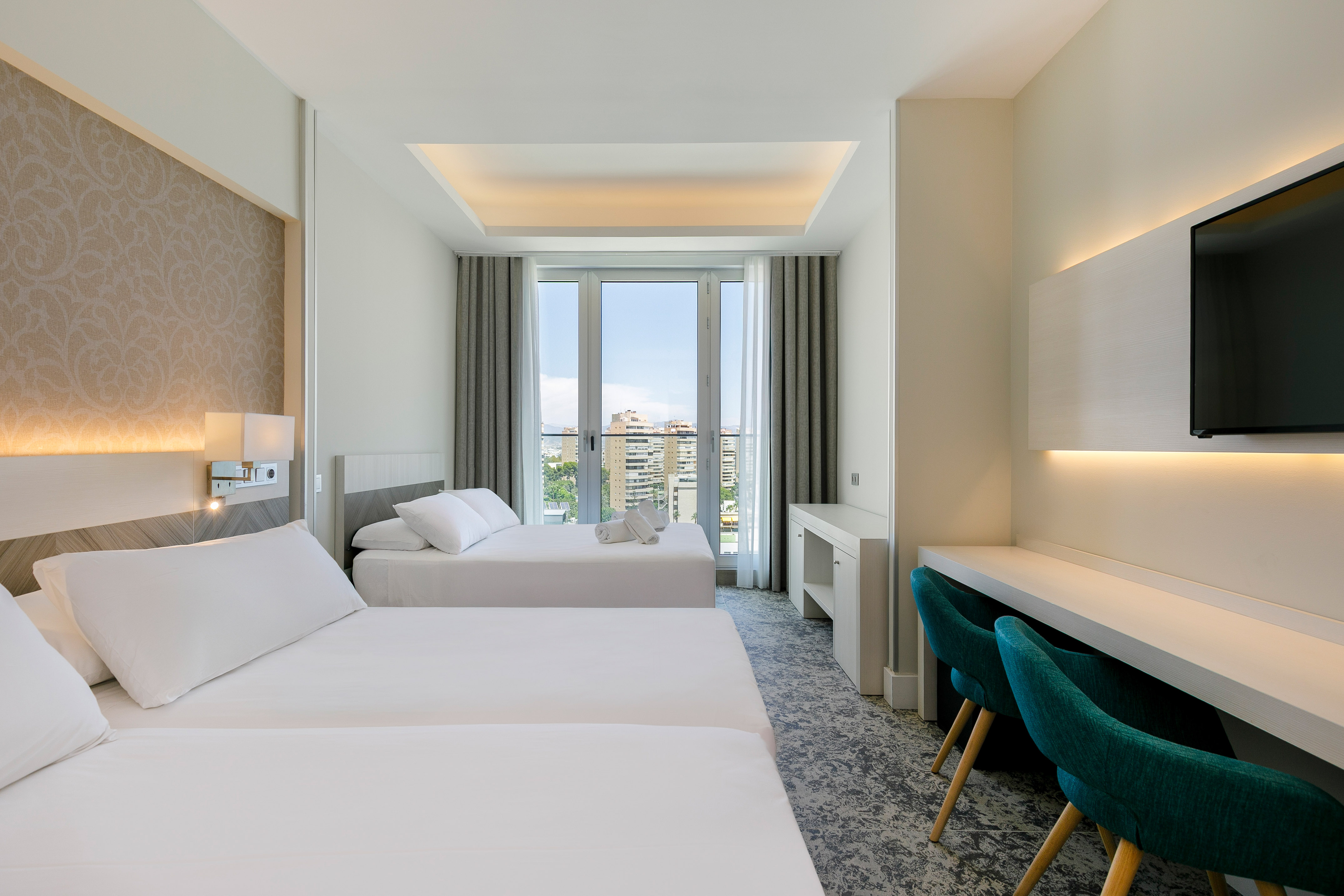 Modern hotel room with three beds, city view balcony, and desk area.