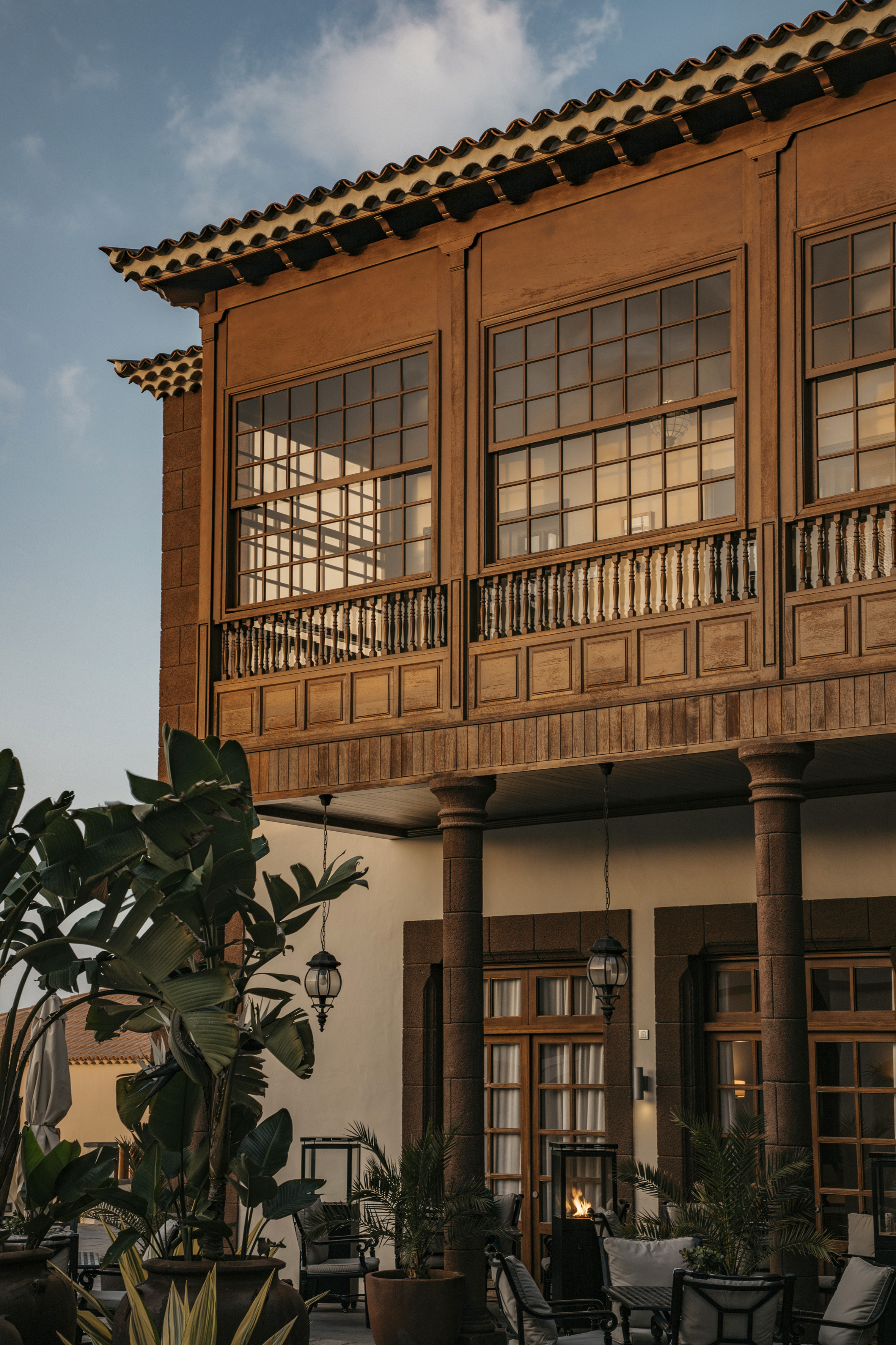 Wooden building facade with large windows, balcony, tiled roof, and lush plants.