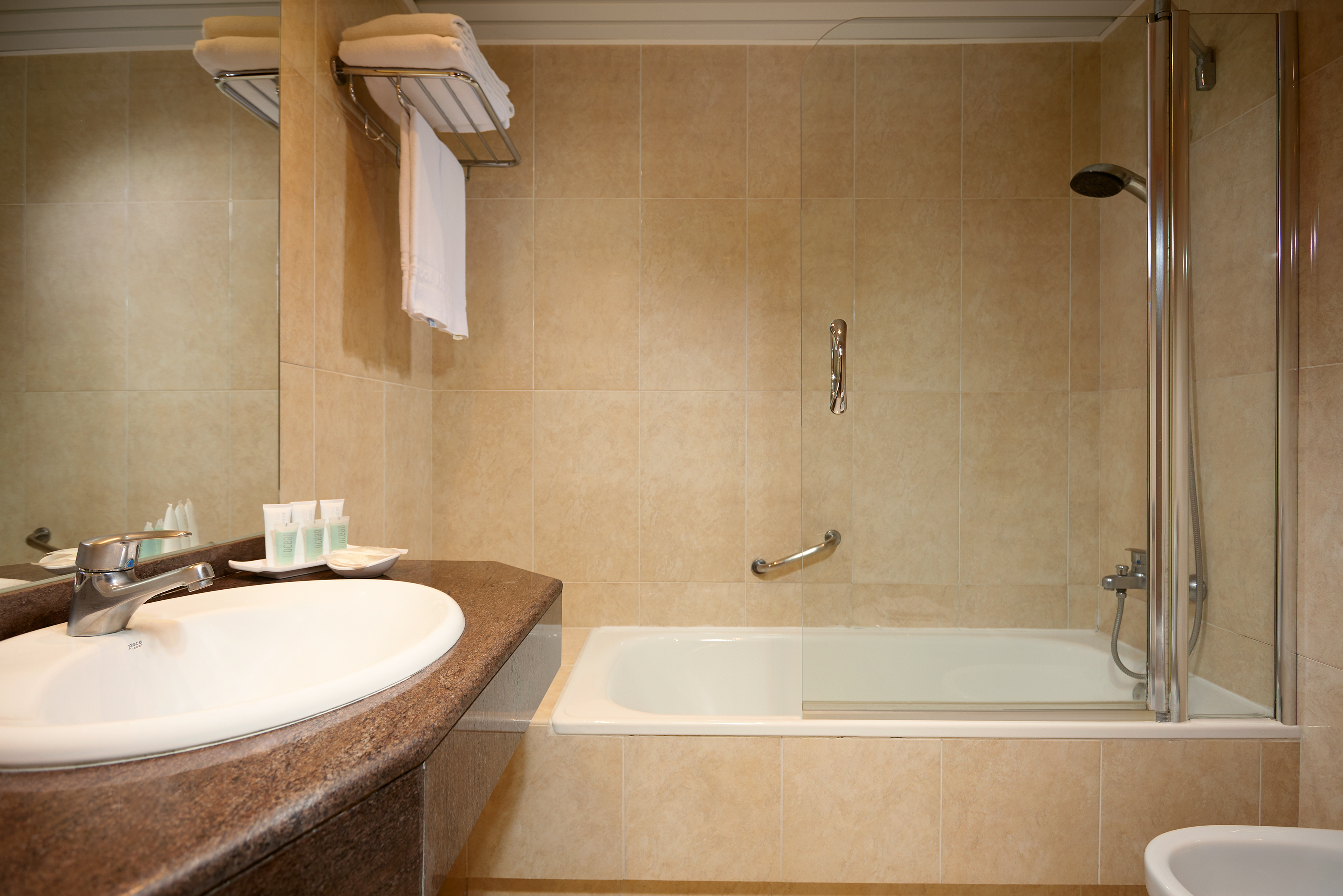 Clean hotel bathroom with beige tiles, bathtub, shower, sink, and fresh towels.