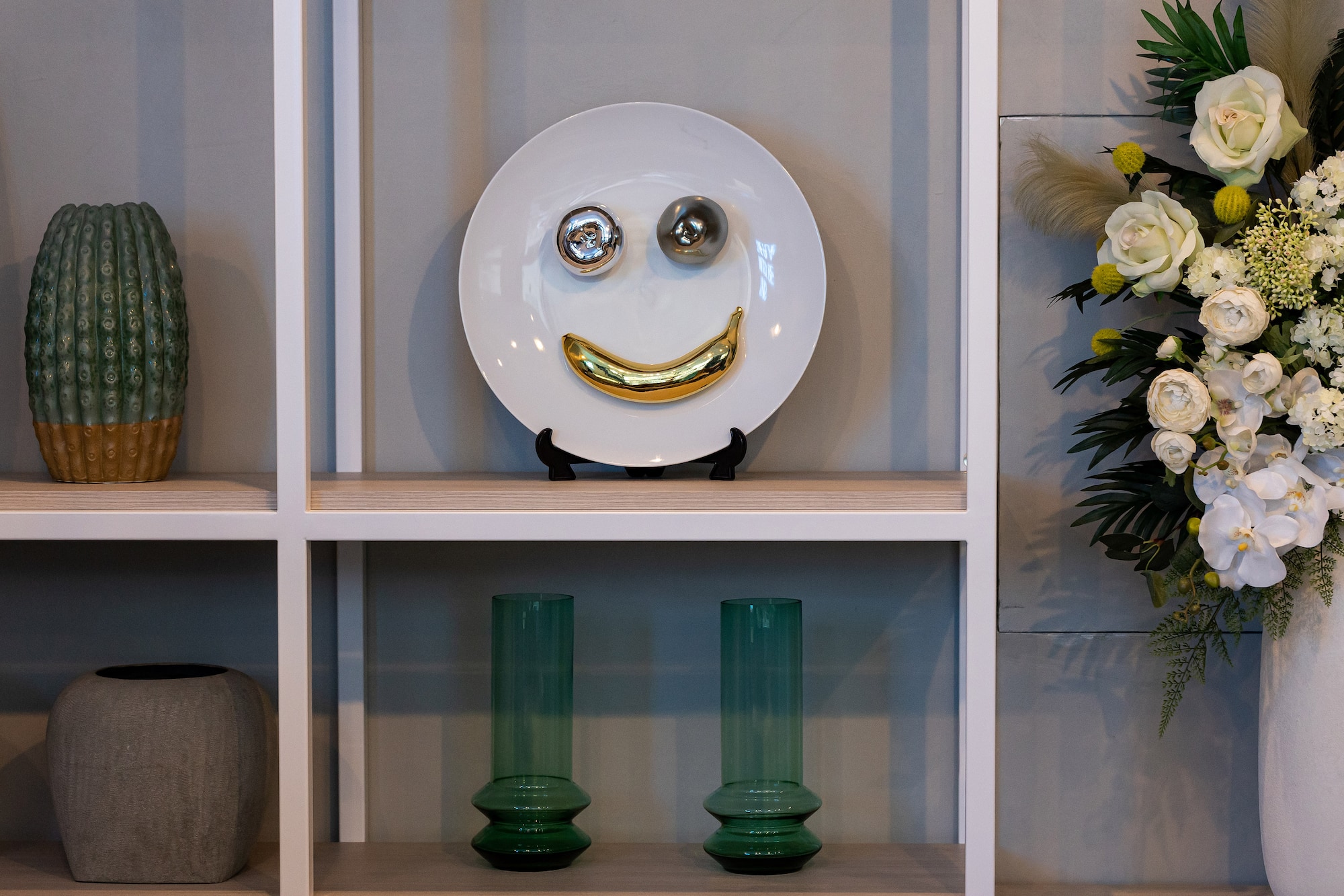 Modern home decor: white plate with gold banana smile, silver eyes on shelf.