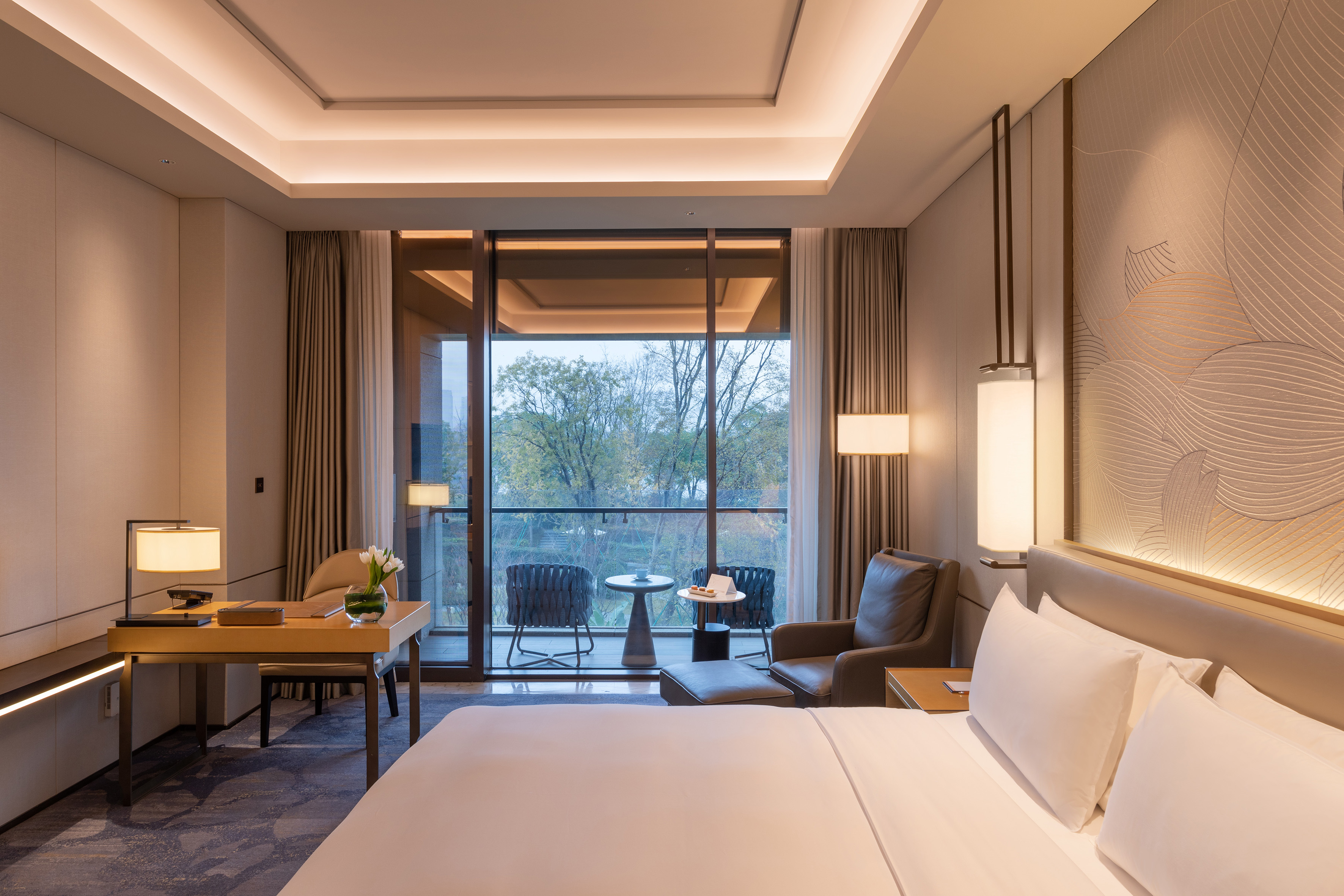 Modern hotel room with bed, desk, armchair, and balcony view.