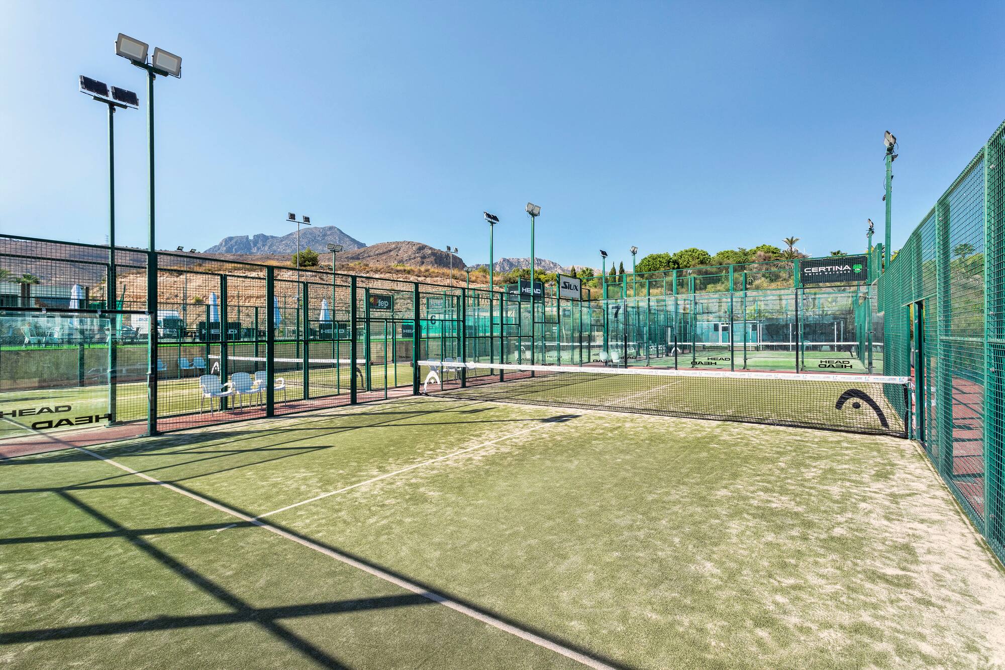 Multiple outdoor padel courts with green turf, nets, fences, and distant mountains.