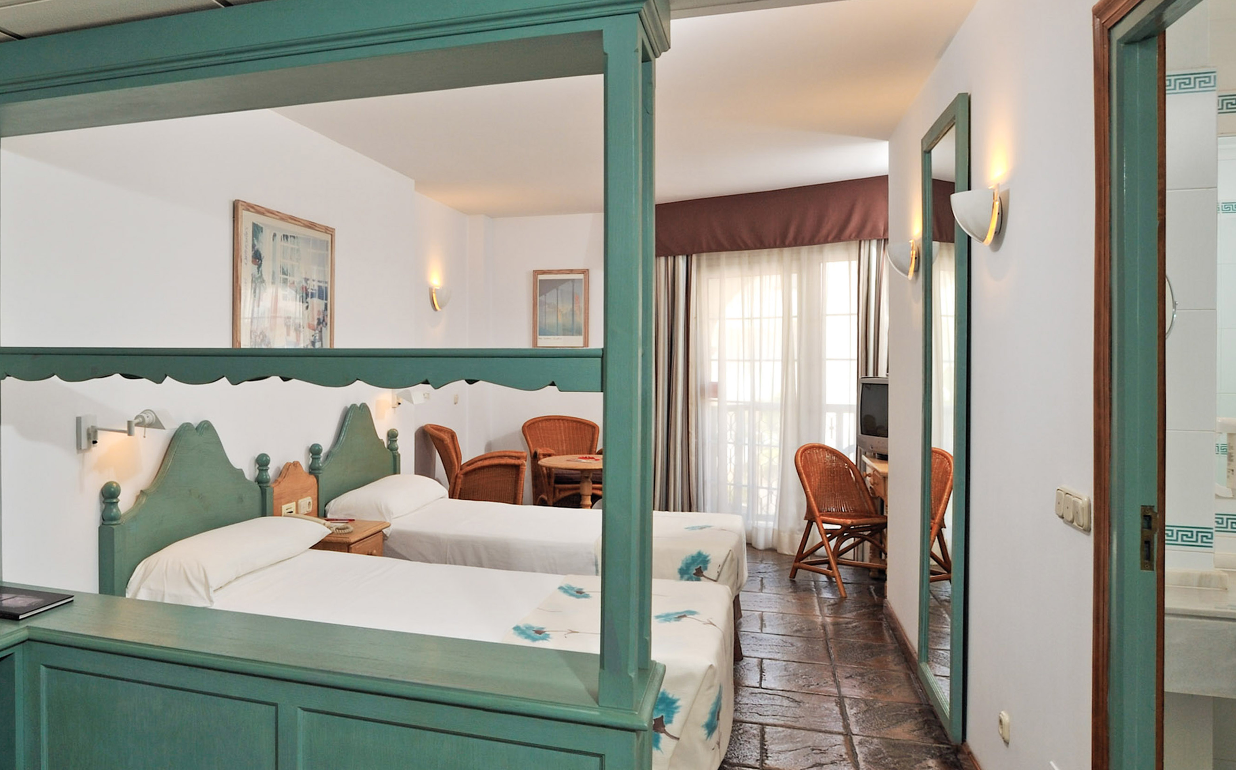 Hotel room with two twin beds, green wooden frames, seating area, and balcony.