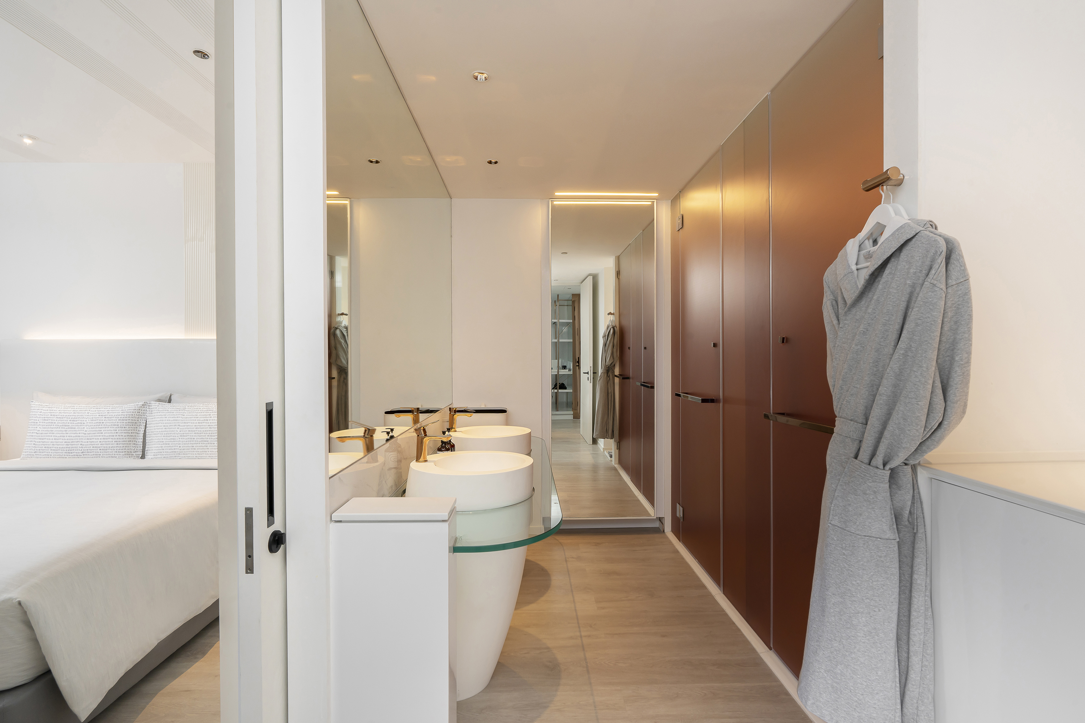 Modern hotel bathroom with double sinks, gold faucets, and attached bedroom.