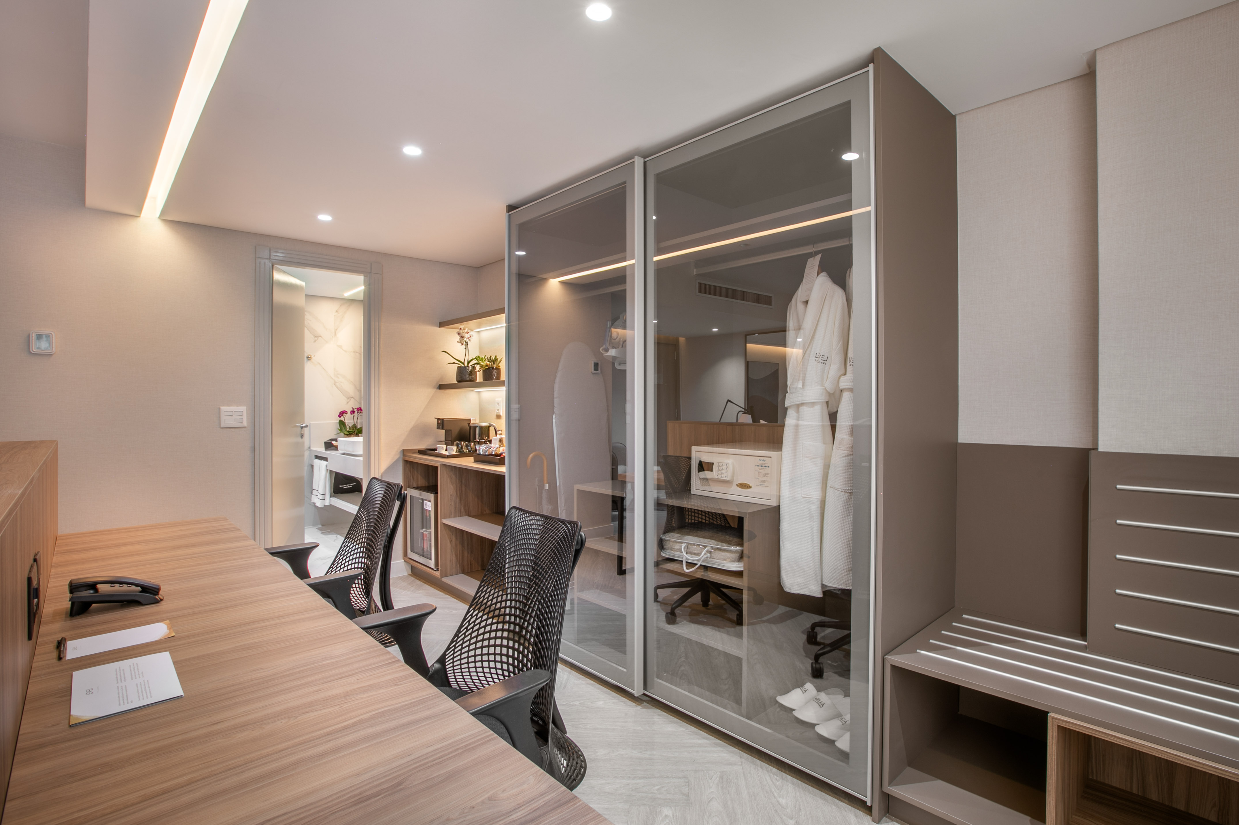 Modern hotel room workspace with glass closet, robes, and minibar.