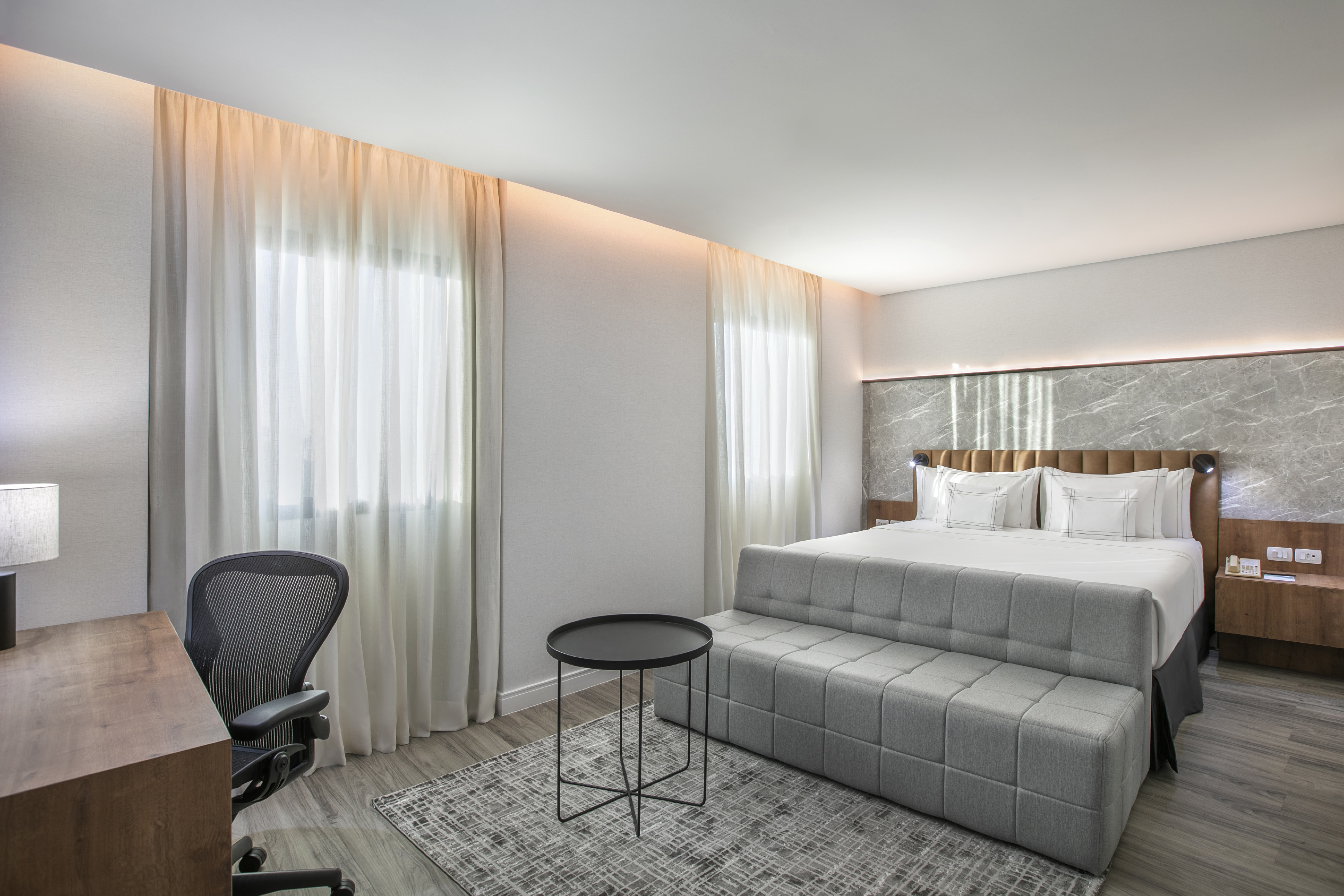 Modern hotel room with king bed, workspace, grey bench, and marble wall.