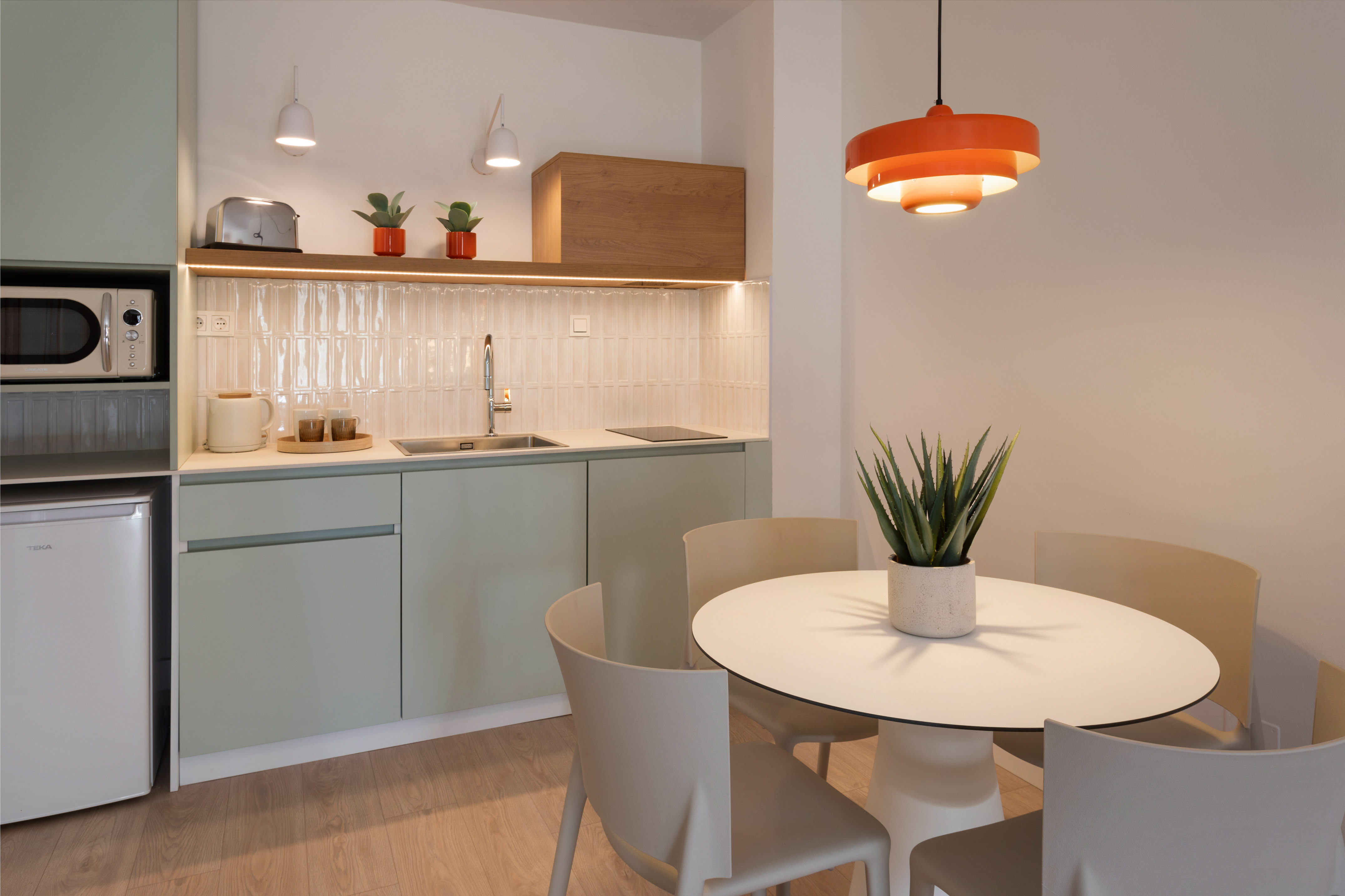 Modern kitchenette with mint green cabinets, white dining table, orange light.