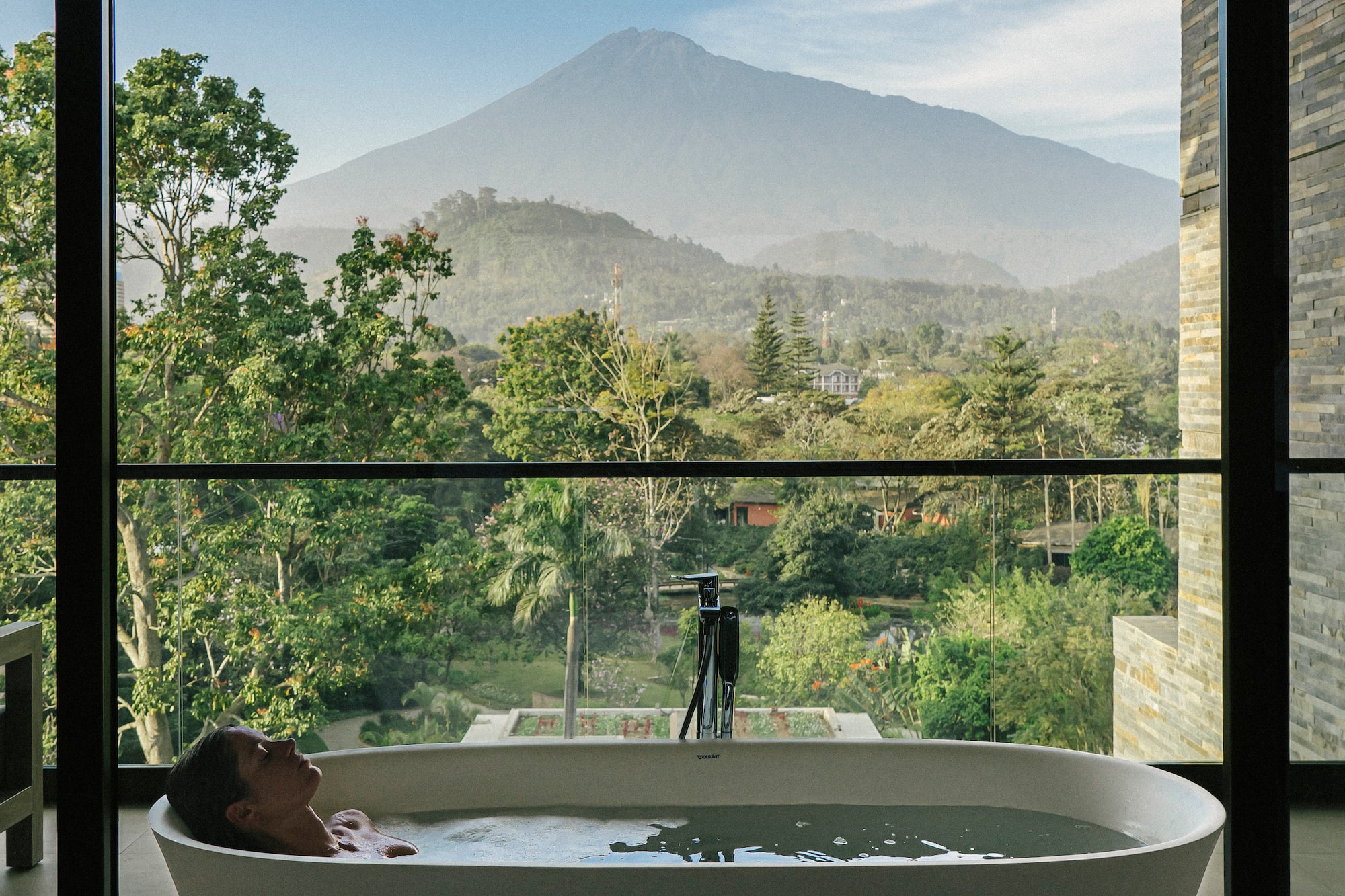 Woman relaxing in bathtub with volcano and green valley view.
