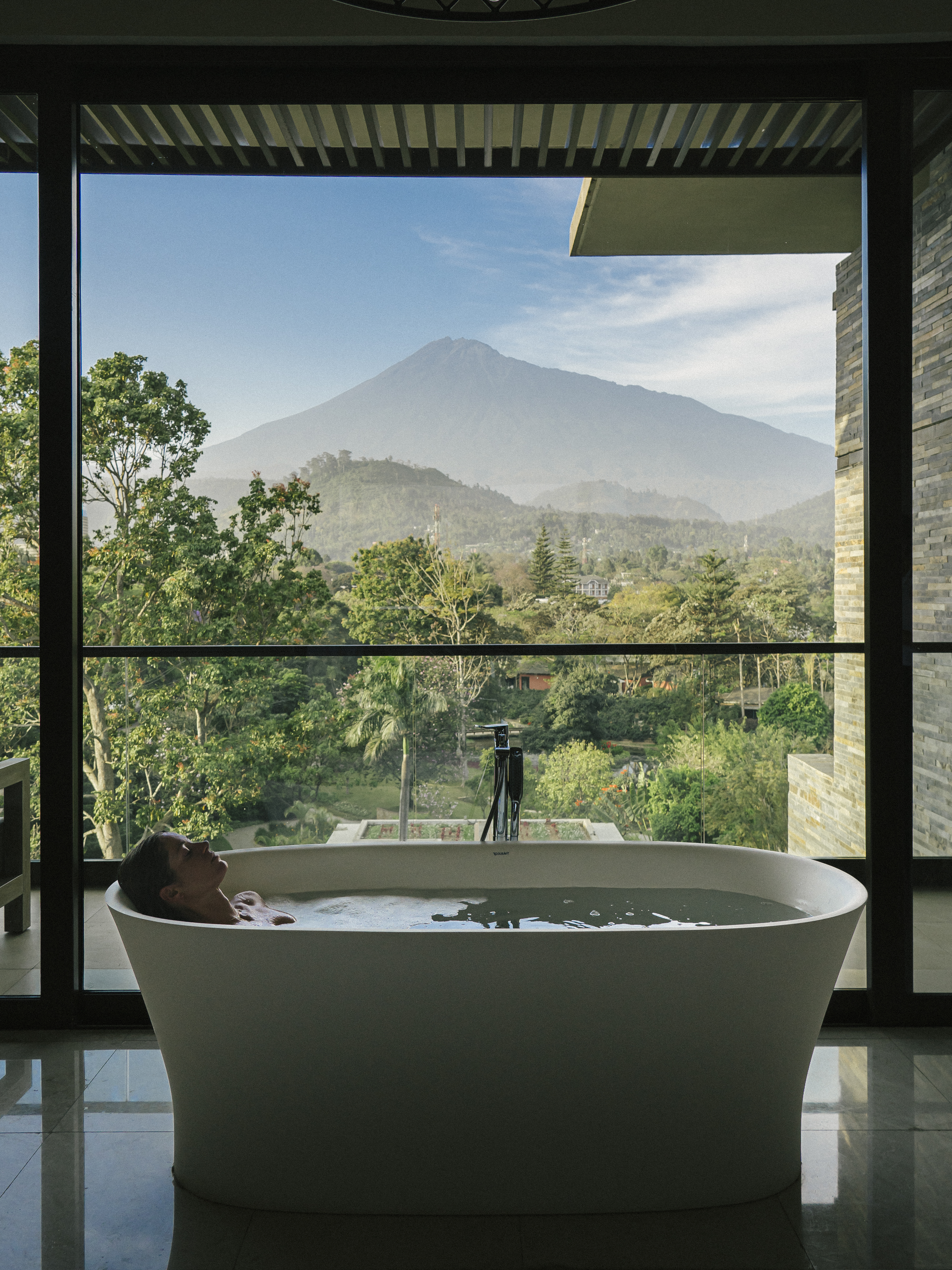 Woman relaxing in bathtub with volcano and green valley view.