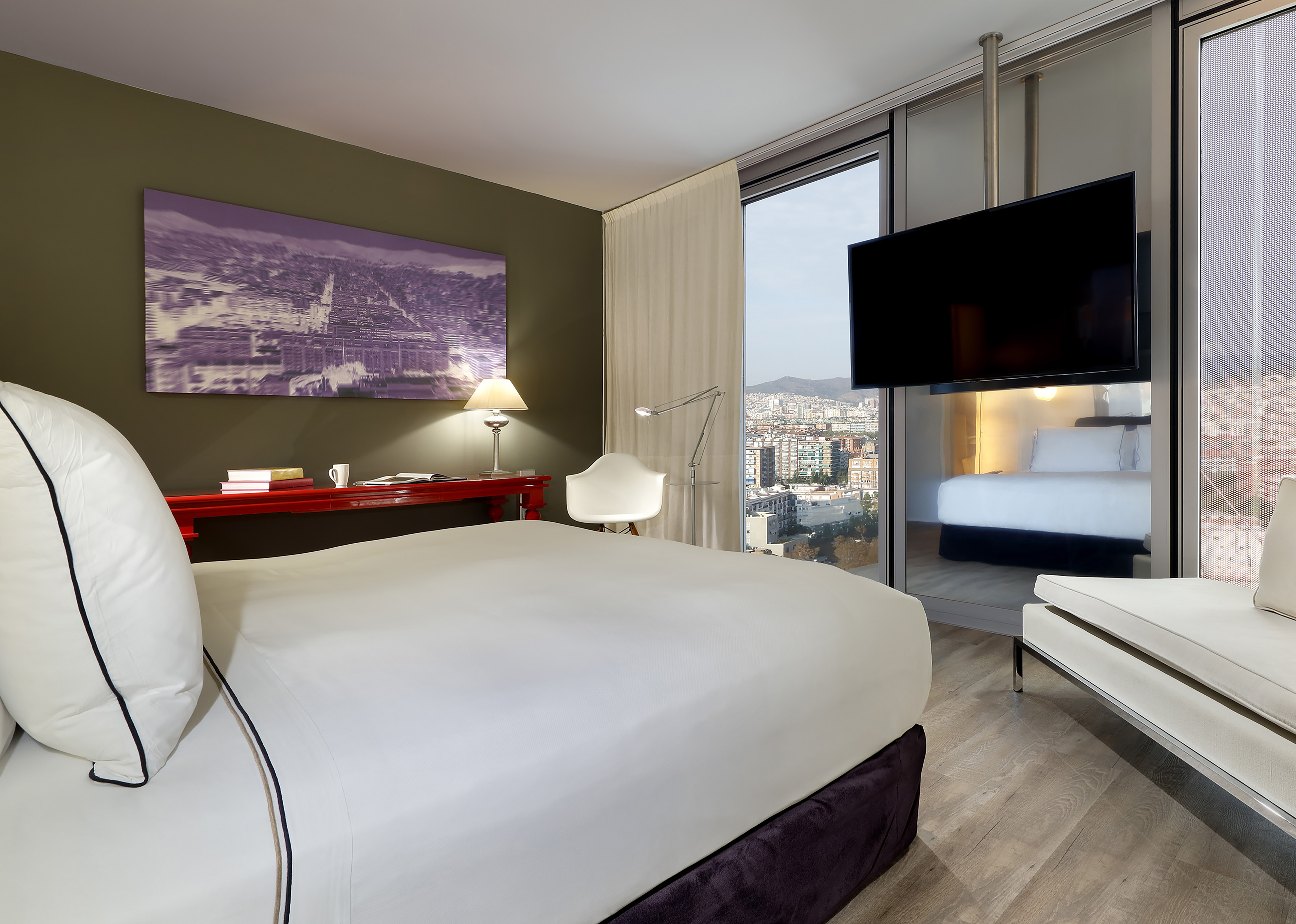 Modern hotel bedroom with city view, large bed, red desk, and hanging TV.