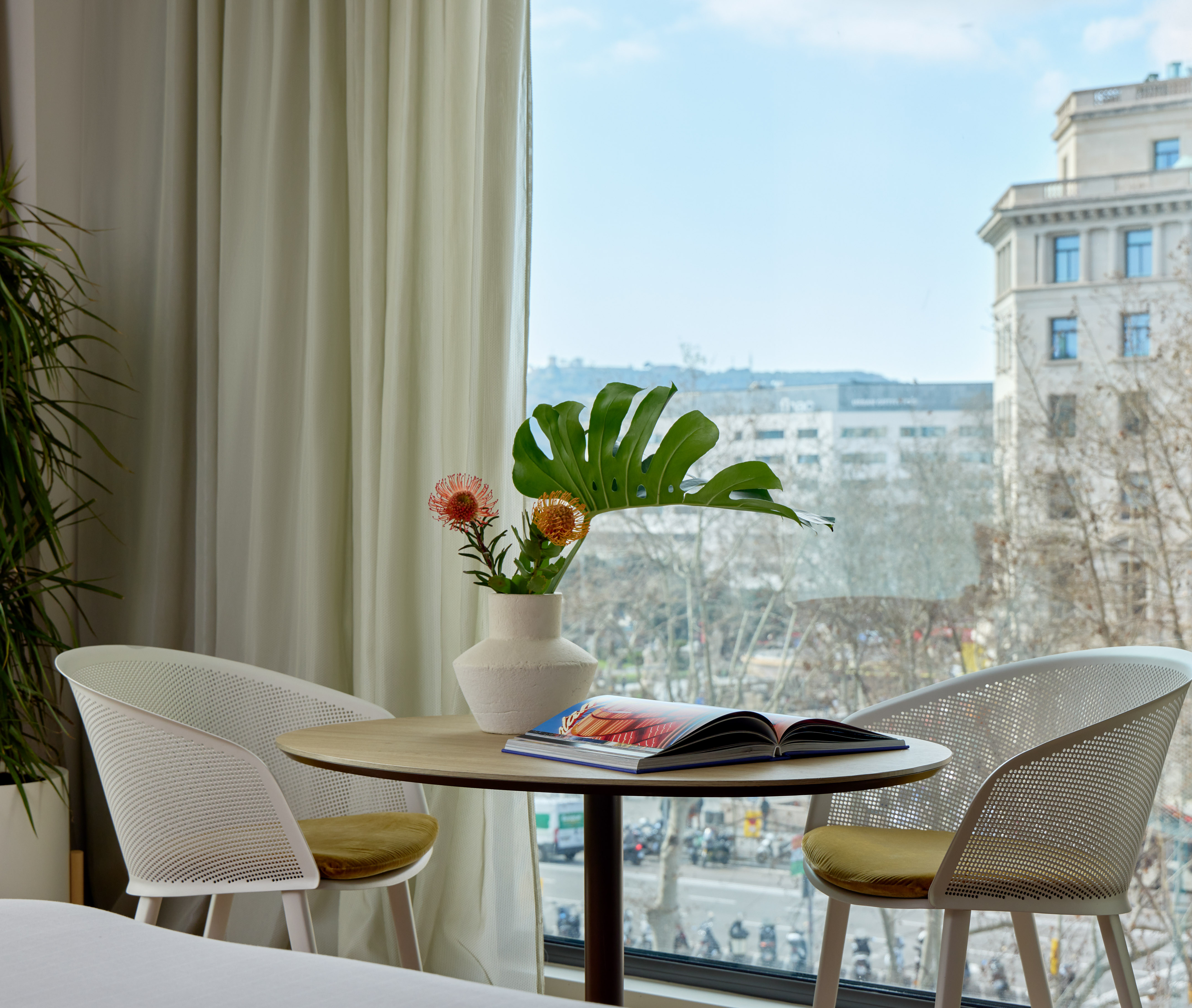 Modern hotel room, city view, table with flowers, open book, and two chairs.