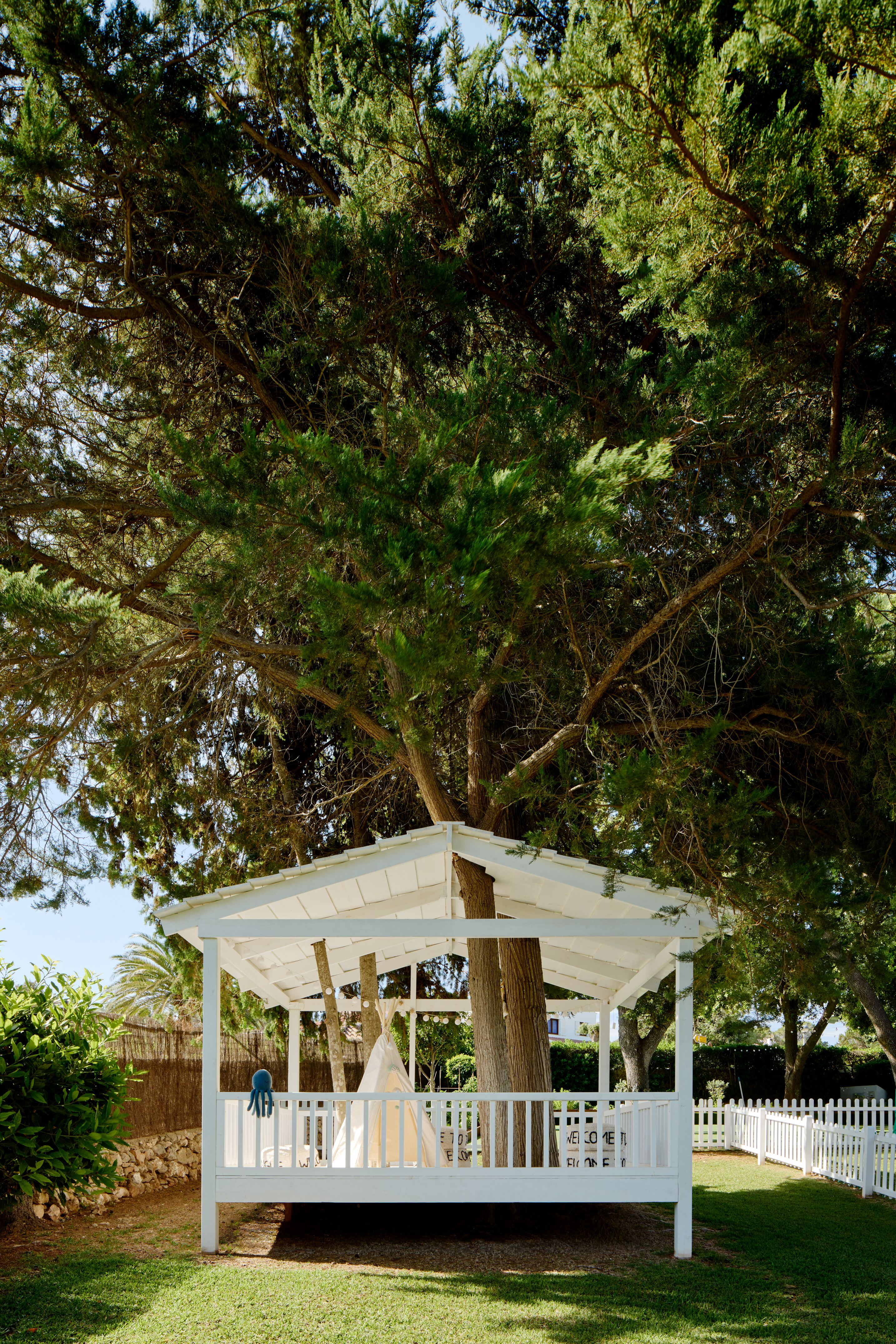 White garden playhouse with teepee and octopus toy around a large tree.