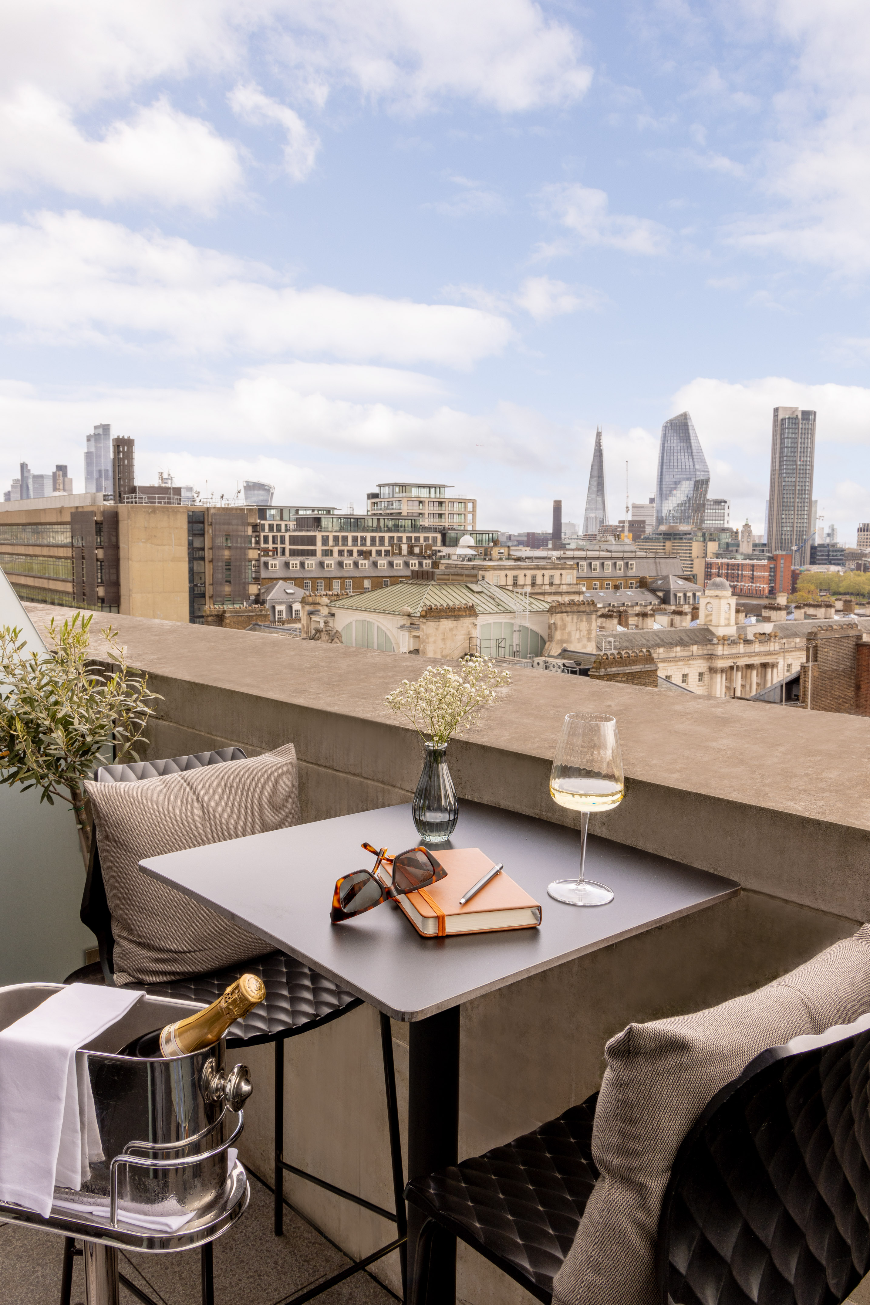 A balcony with a table holding glasses and books, framed by a city skyline in the background.