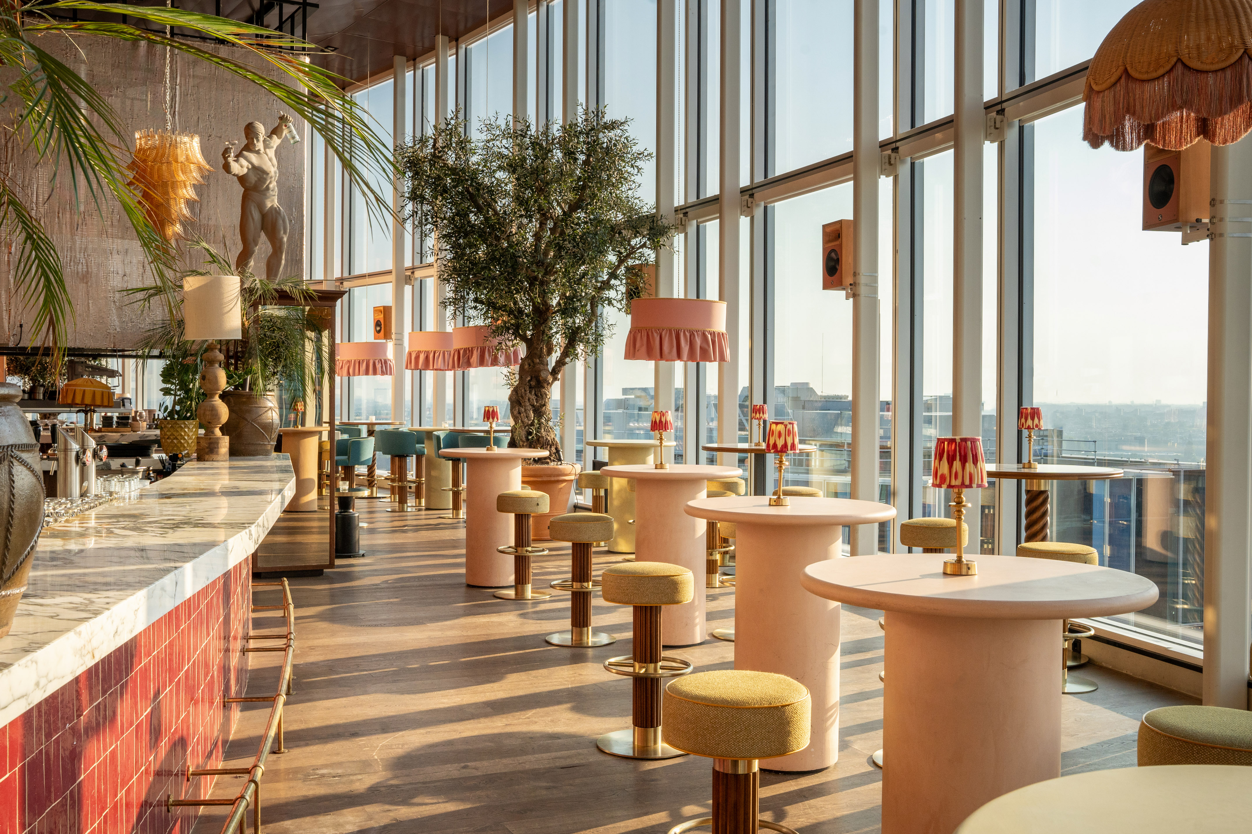Modern rooftop bar with marble counter, pink tables, olive tree, city skyline.