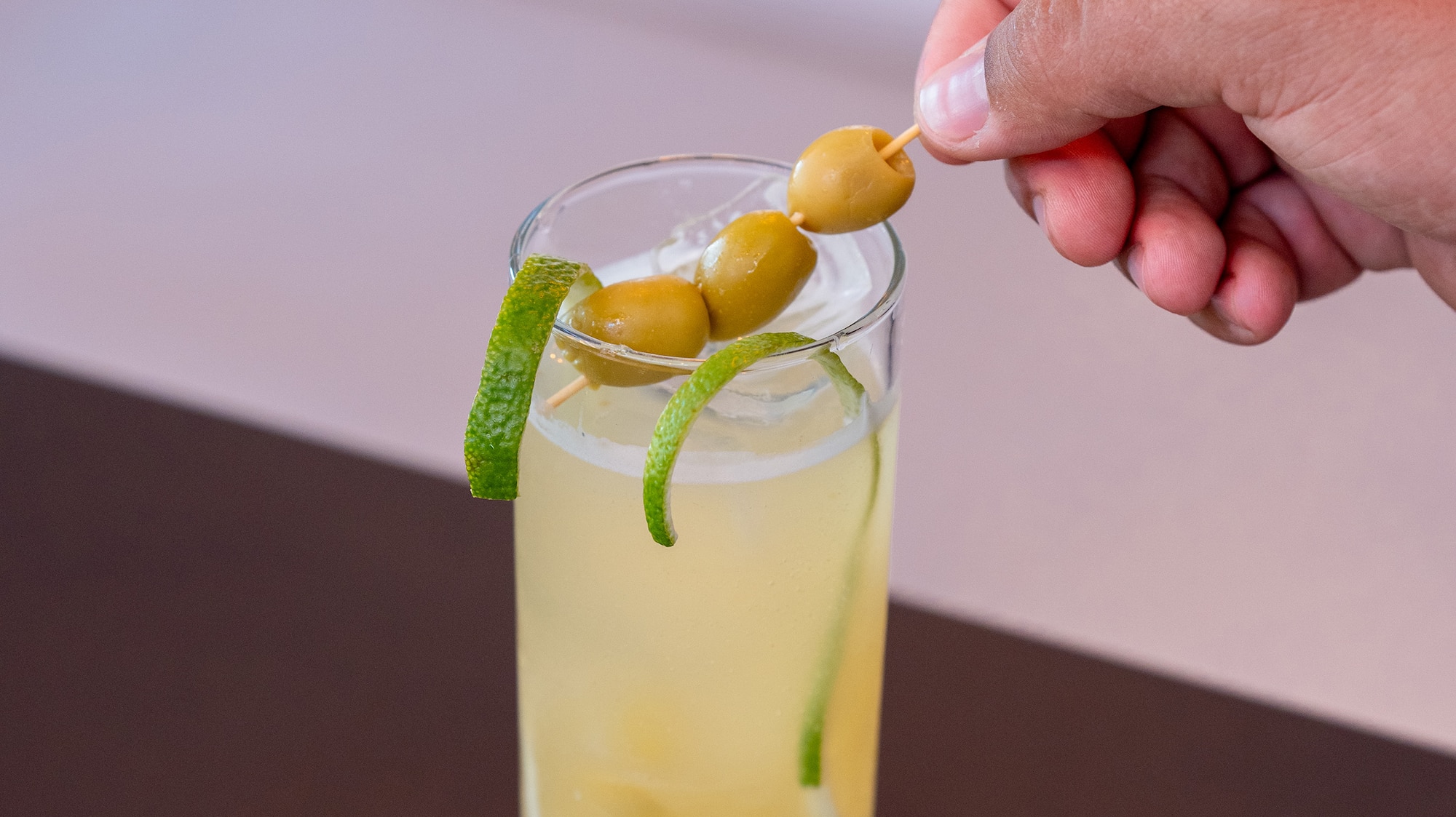 a hand holding a stick with olives on a stick in a glass of liquid