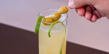 a hand holding a stick with olives on a stick in a glass of liquid