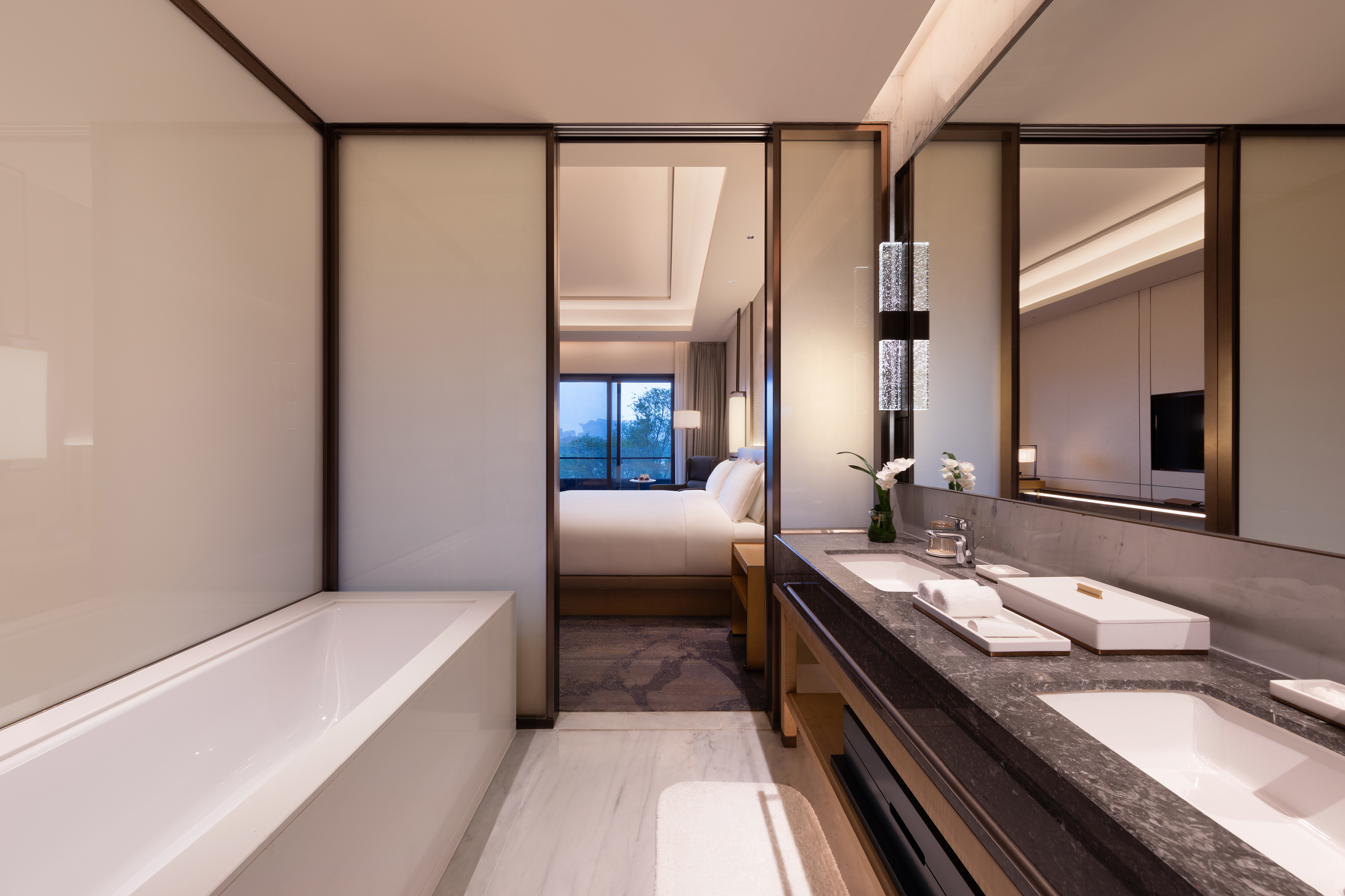 Modern hotel bathroom with bathtub, double vanity, opening to luxurious bedroom.
