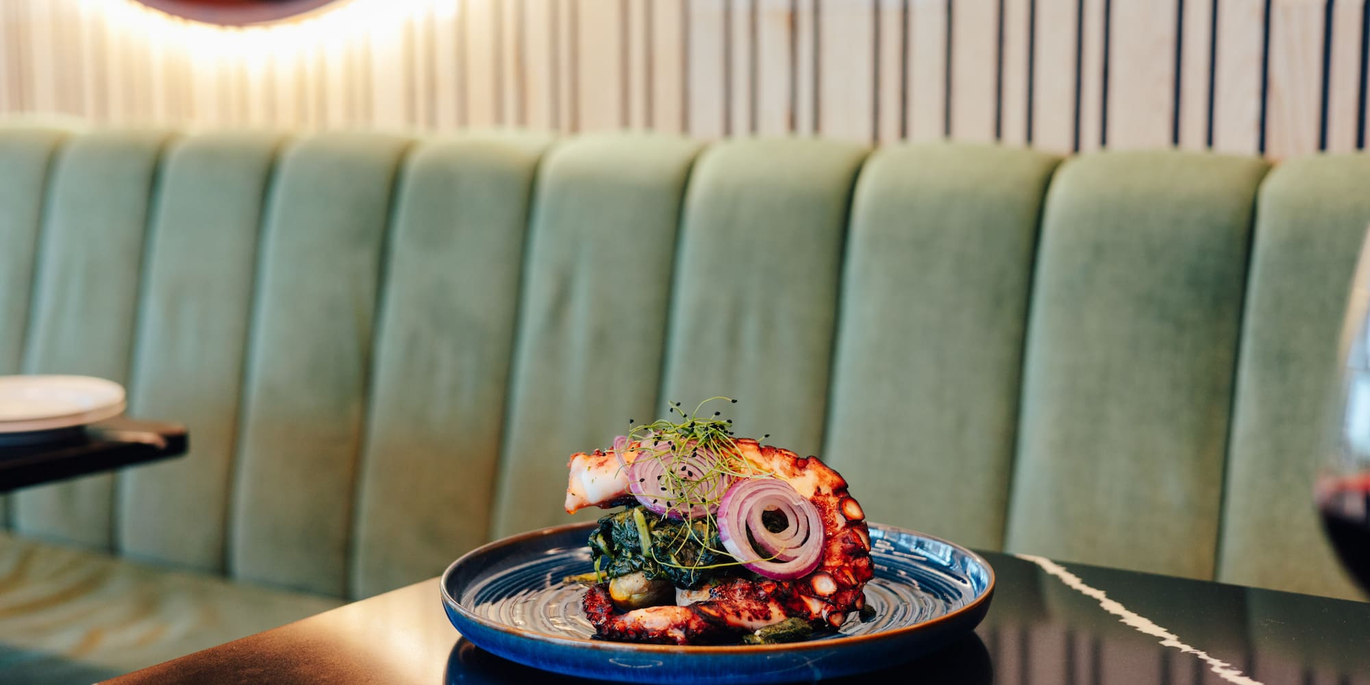 Grilled octopus seafood with red onion, microgreens on blue plate, restaurant setting.