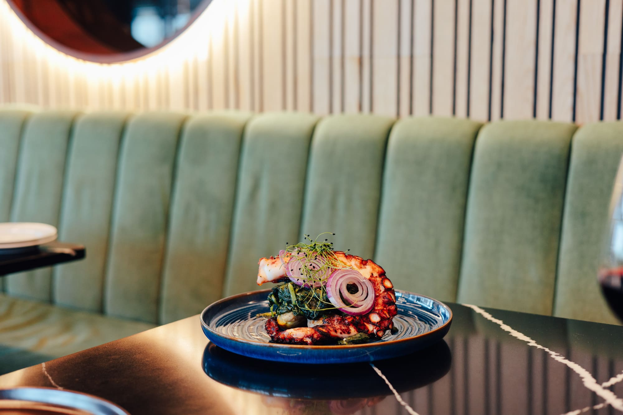 Grilled octopus seafood with red onion, microgreens on blue plate, restaurant setting.