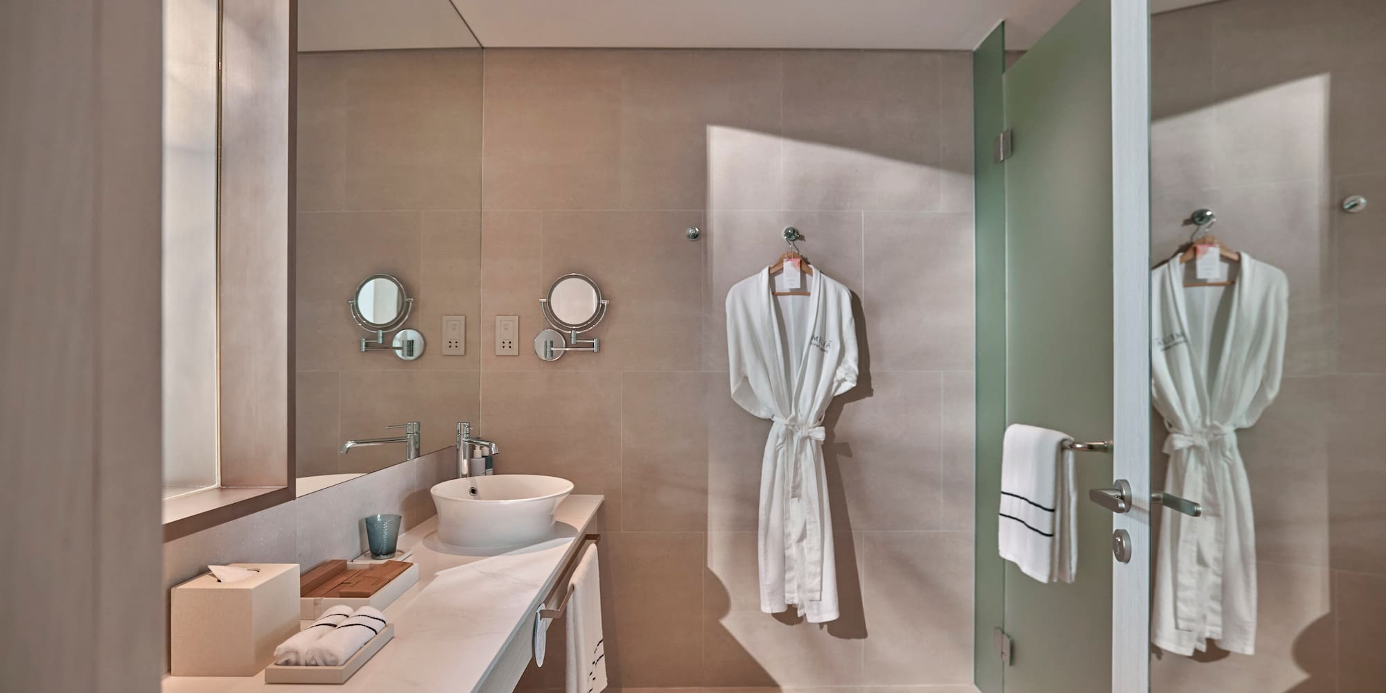 Modern hotel bathroom with vessel sink, beige walls, white robes, glass shower.
