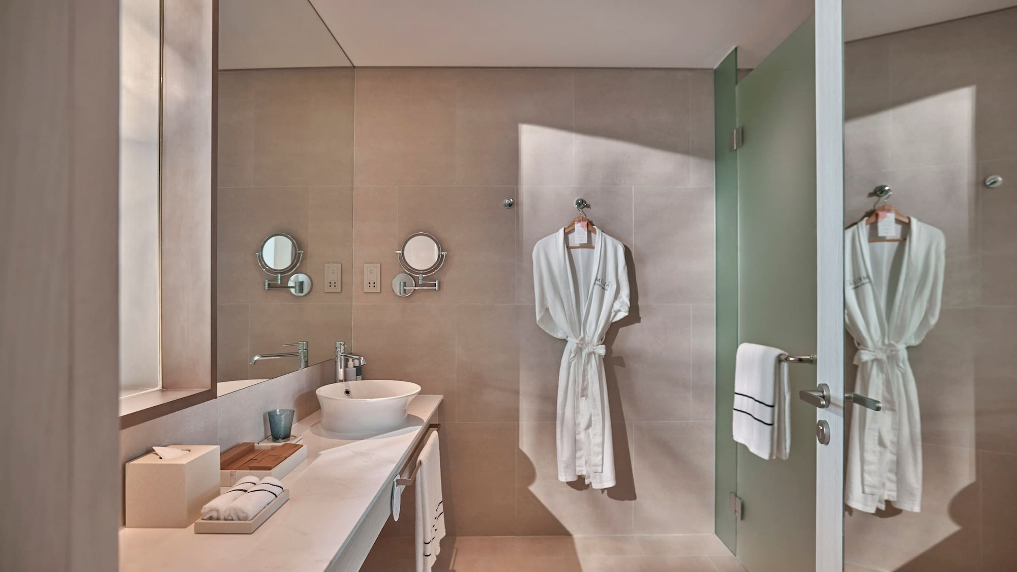 Modern hotel bathroom with vessel sink, beige walls, white robes, glass shower.