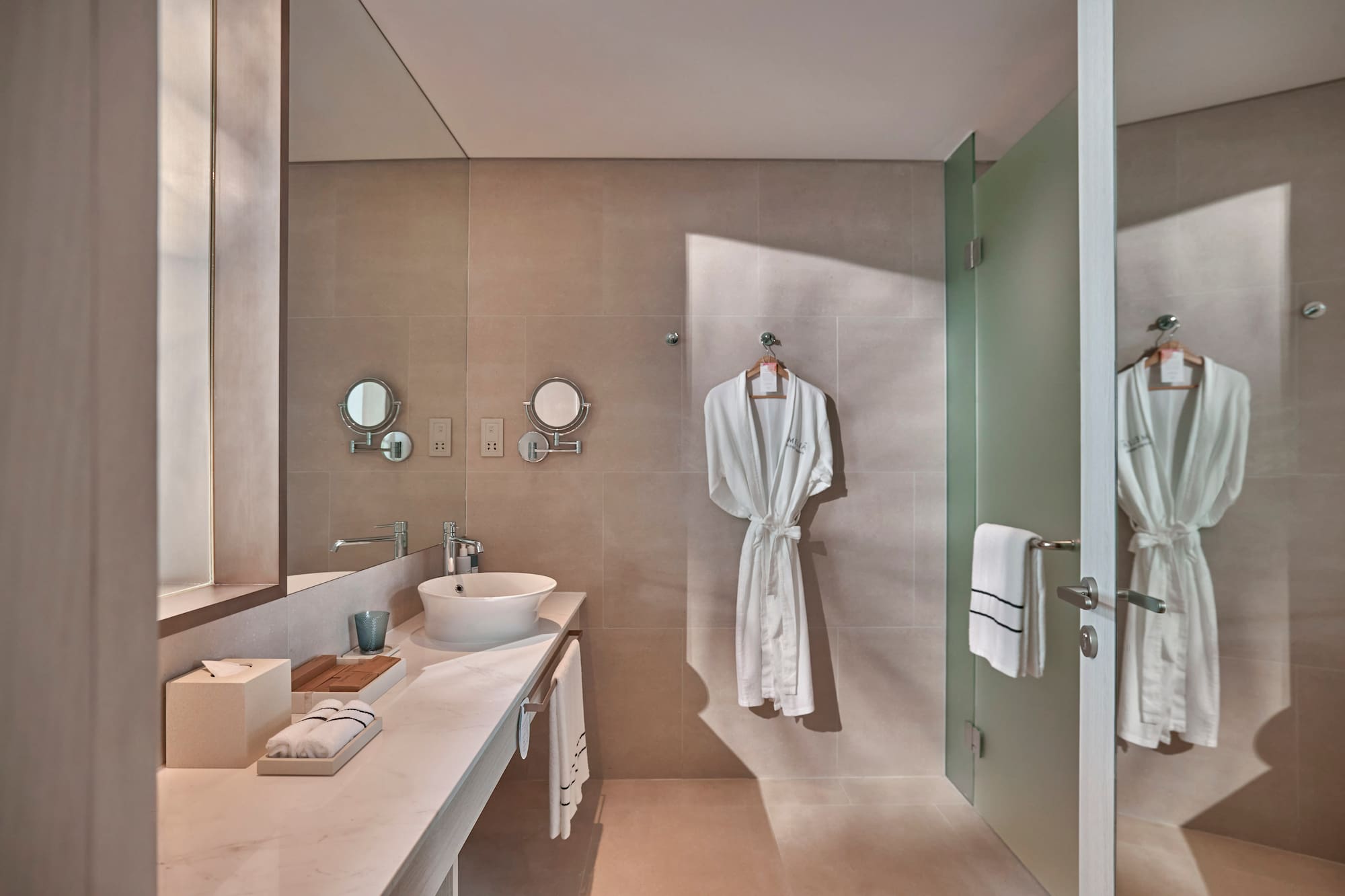 Modern hotel bathroom with vessel sink, beige walls, white robes, glass shower.