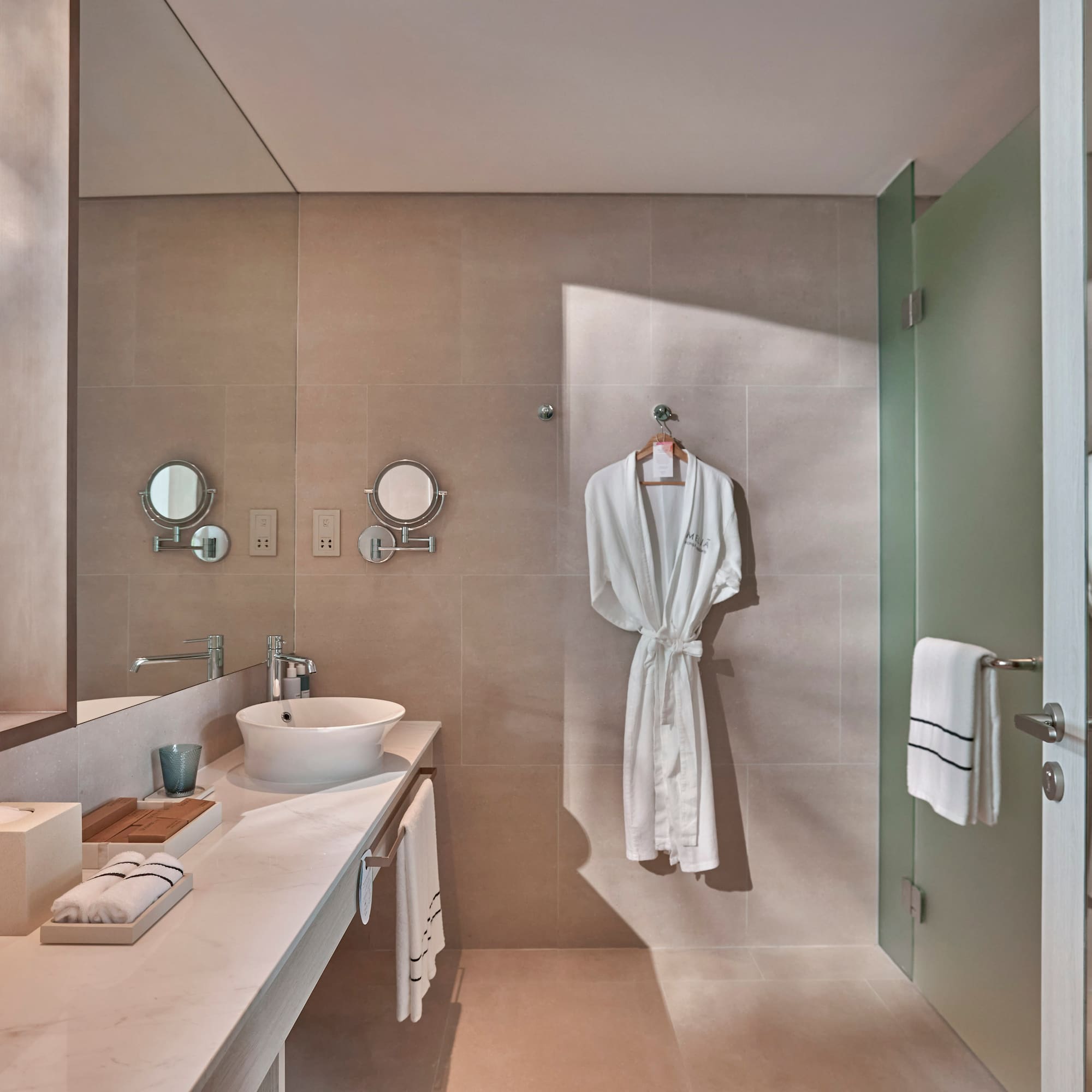Modern hotel bathroom with vessel sink, beige walls, white robes, glass shower.