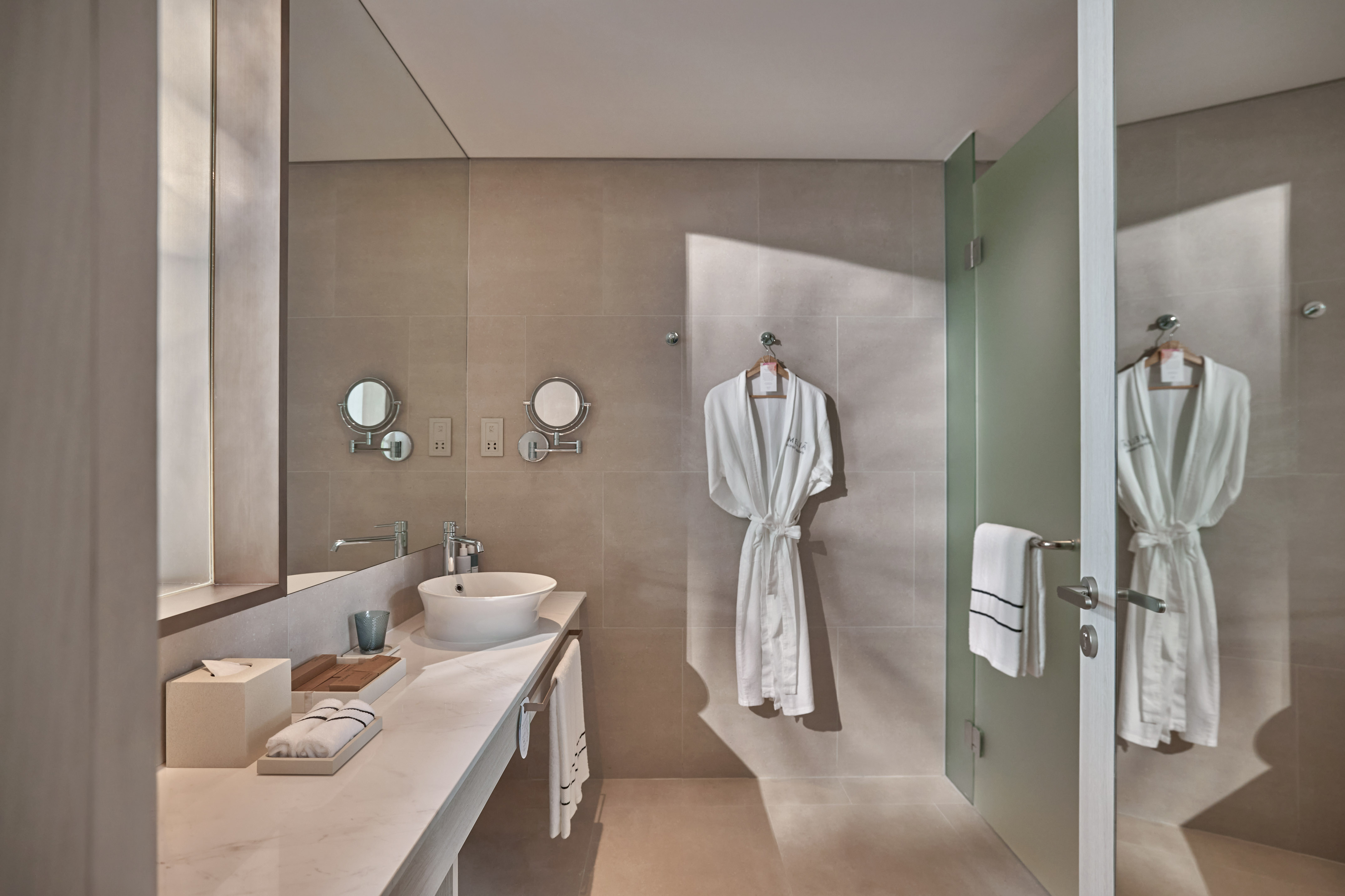 Modern hotel bathroom with vessel sink, beige walls, white robes, glass shower.