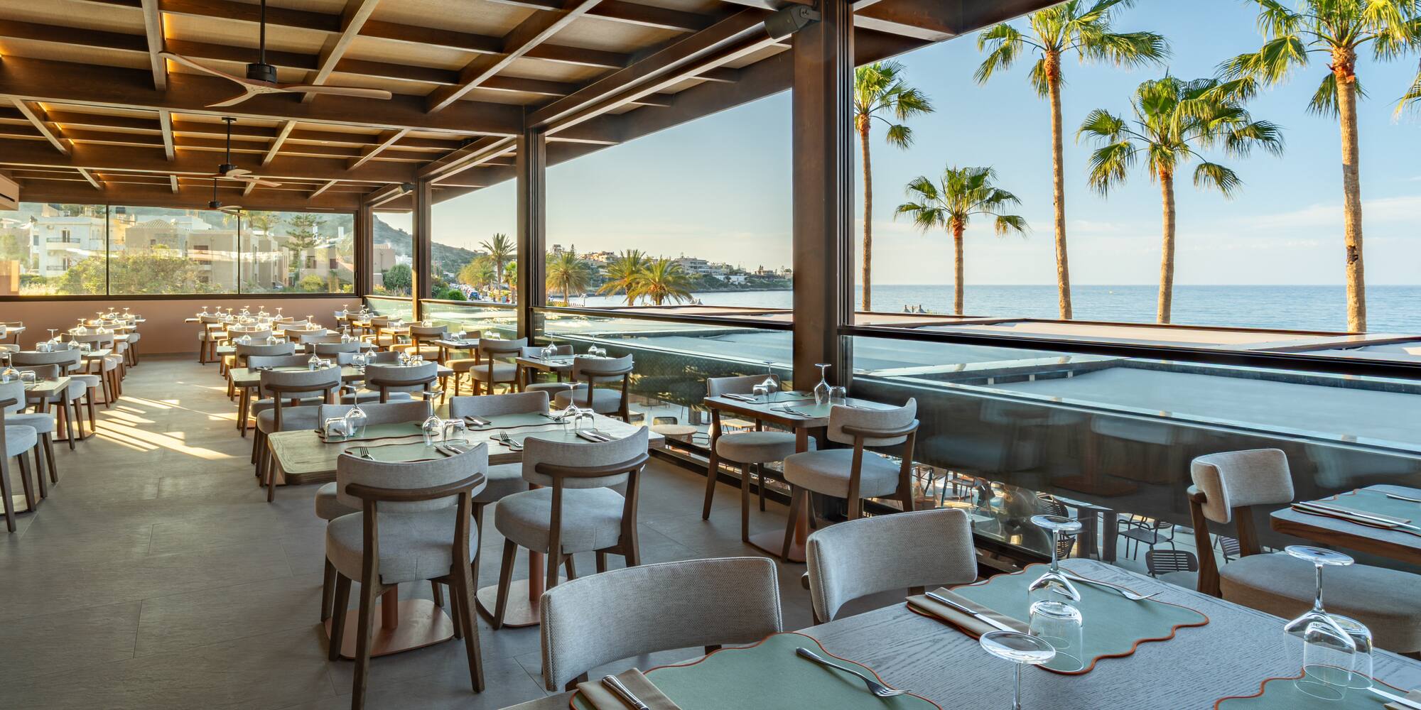 Modern oceanfront restaurant, set tables, palm trees, and sea view.