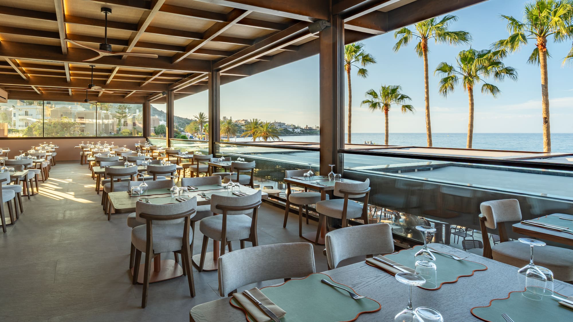 Modern oceanfront restaurant, set tables, palm trees, and sea view.
