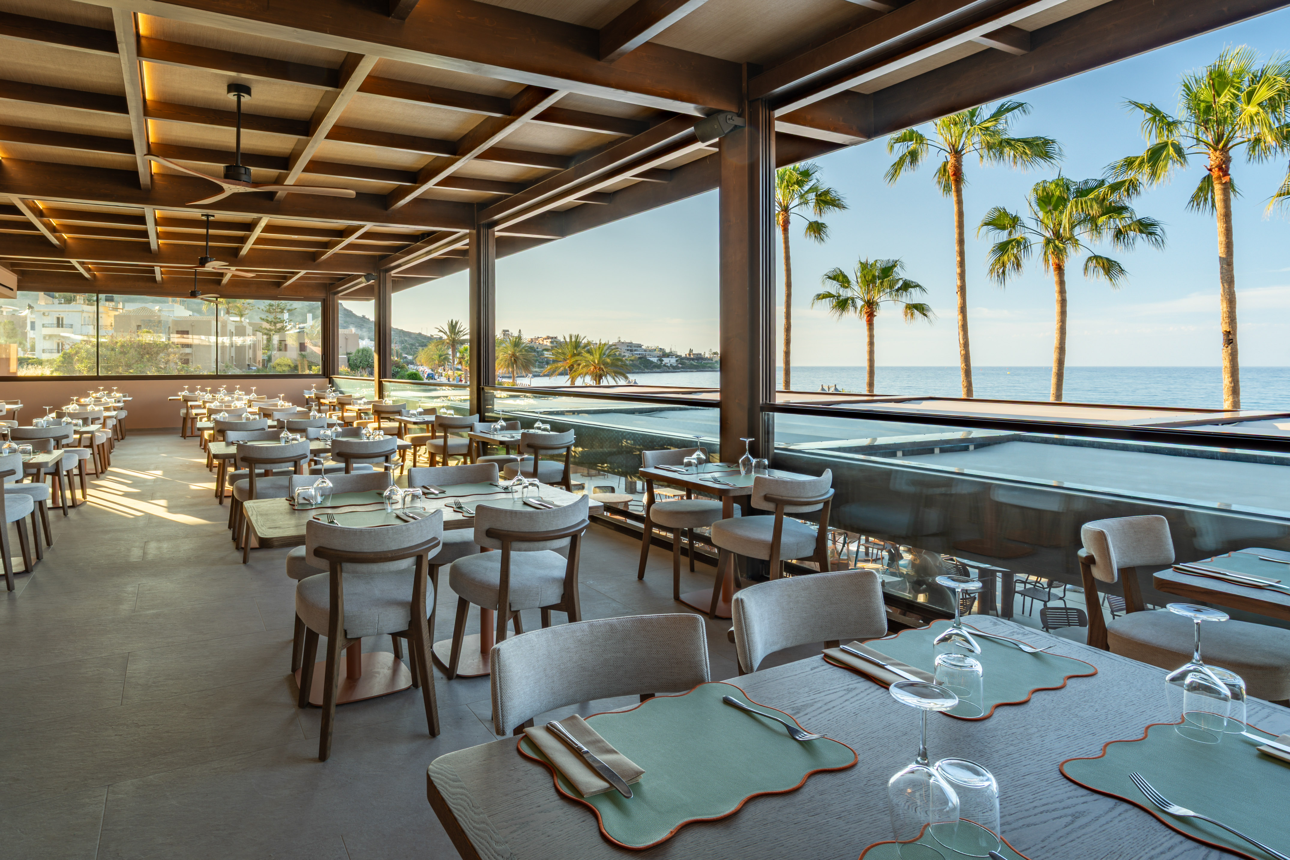 Modern oceanfront restaurant, set tables, palm trees, and sea view.