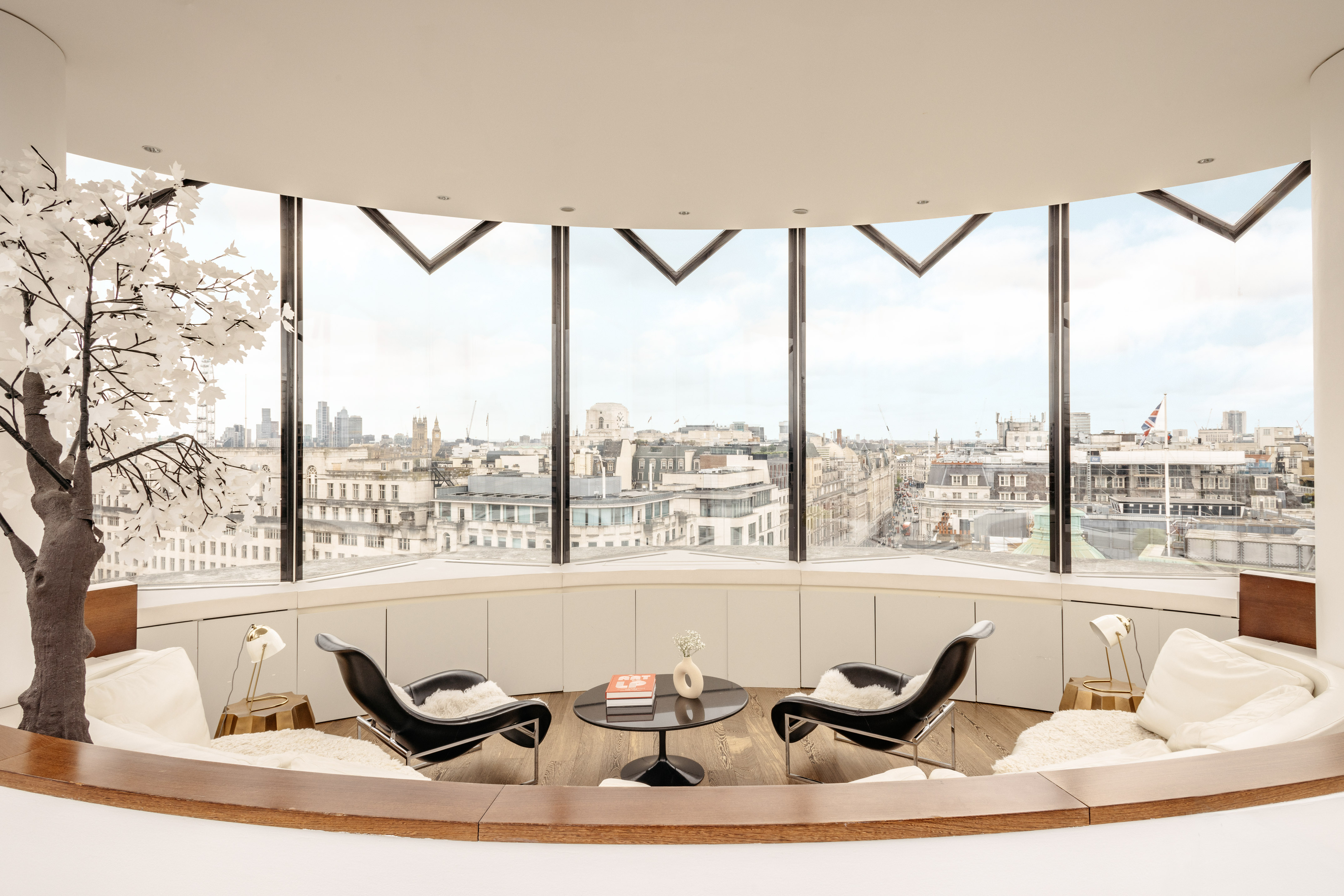 Modern lounge with panoramic London skyline view, decorative tree, and stylish chairs.