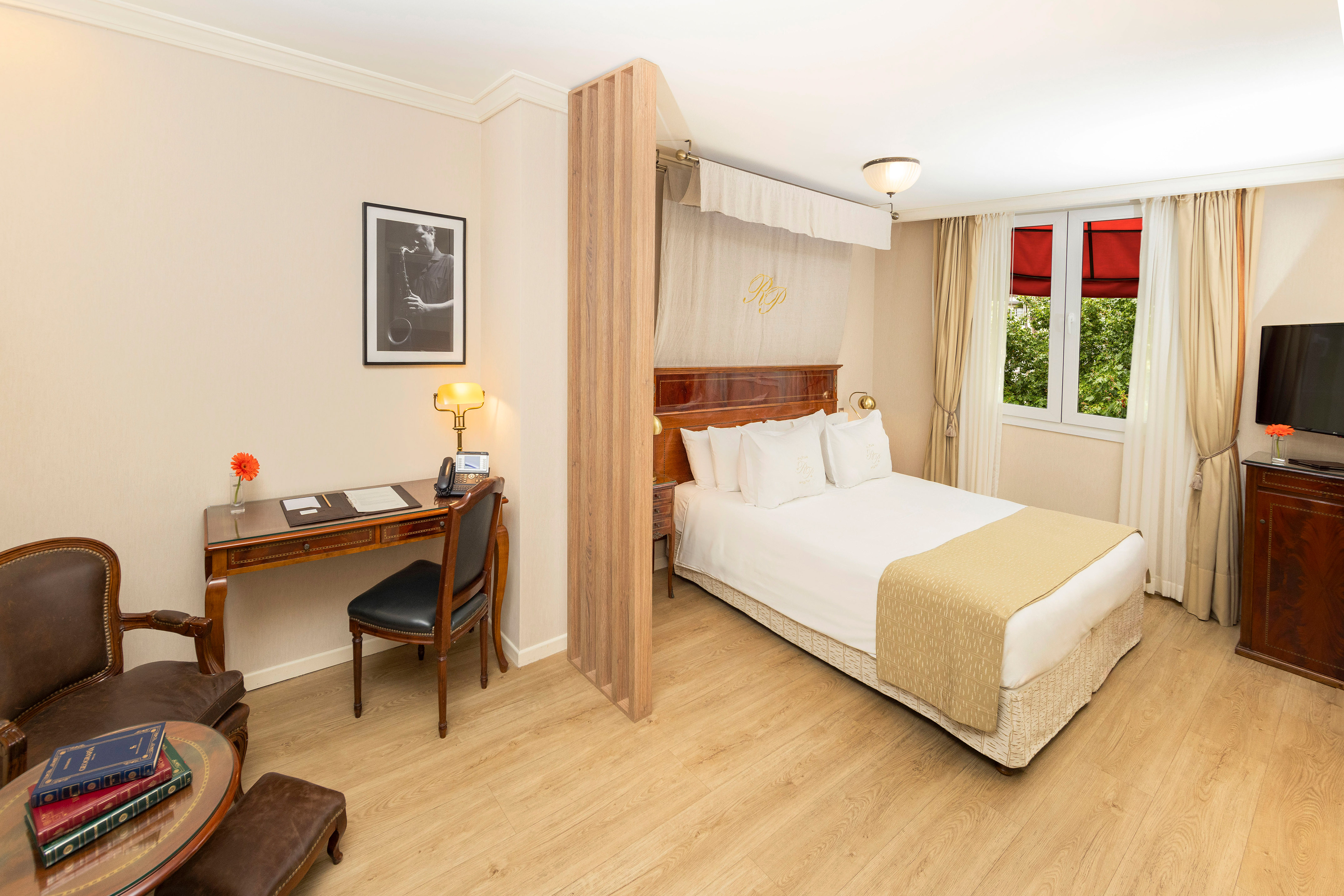 Hotel room with bed, hardwood floors, desk, and sunlit window.