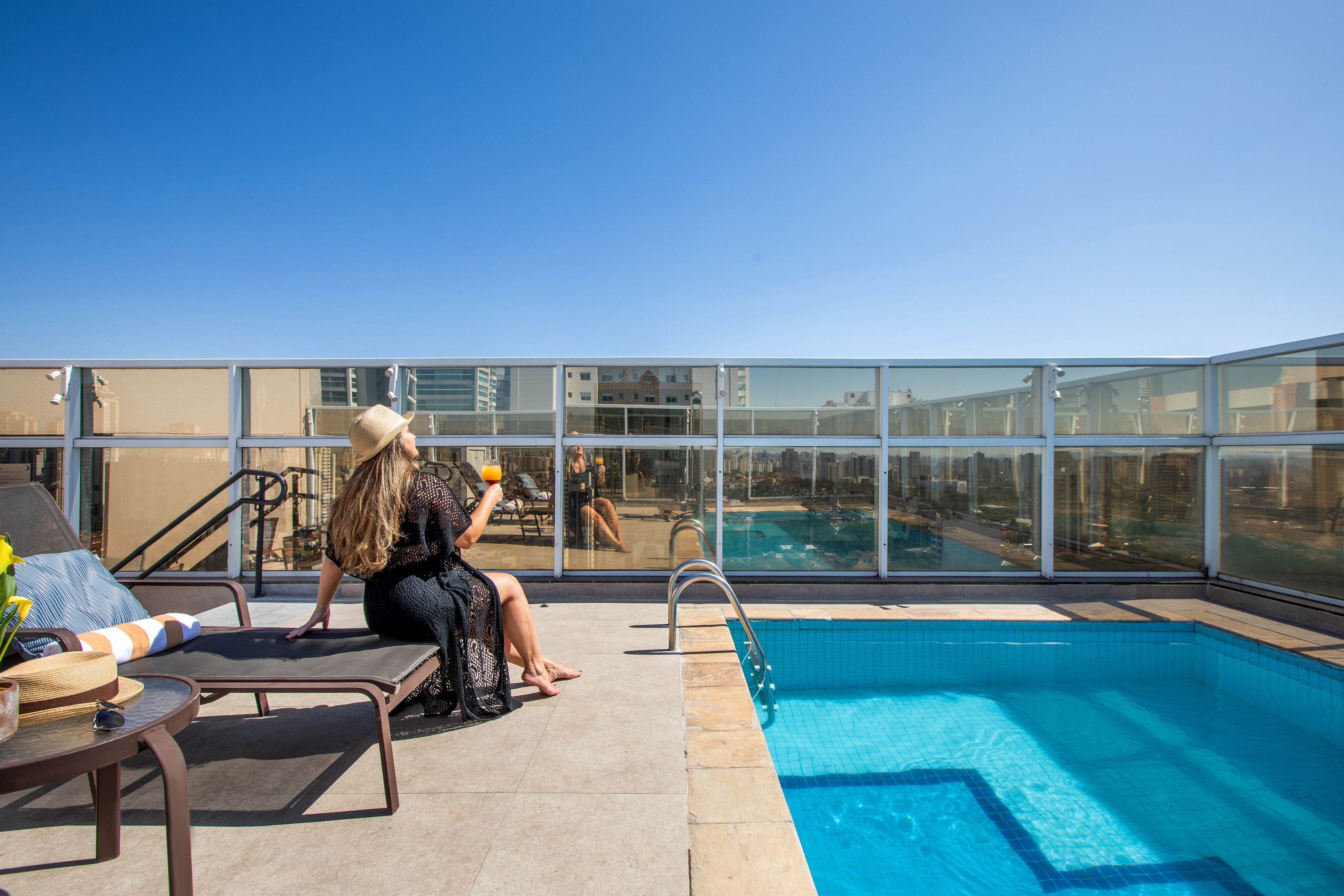 Woman relaxing by rooftop pool with city view under clear sky.