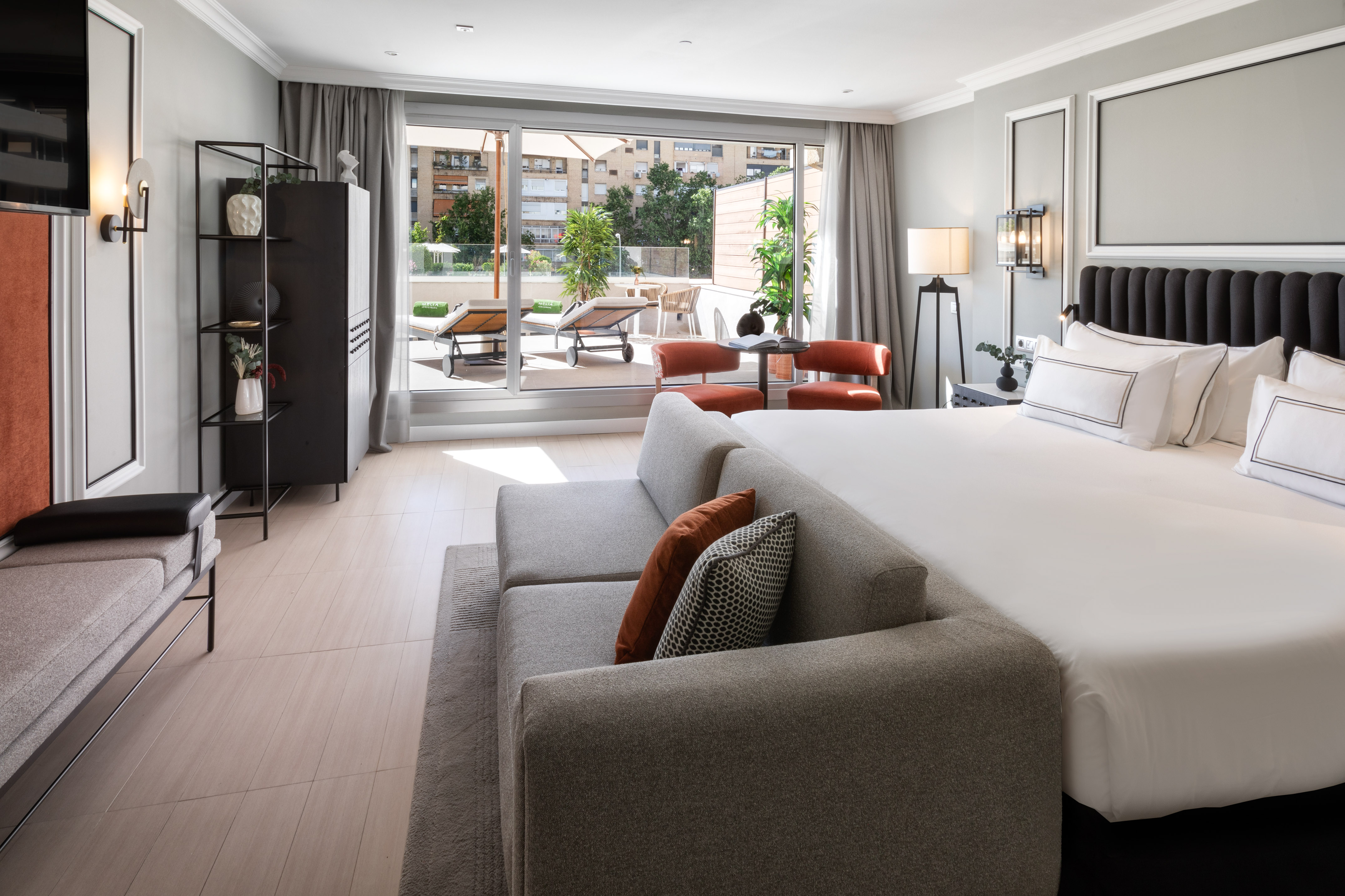 Modern hotel room with king bed, sofa, and private terrace with lounge chairs.