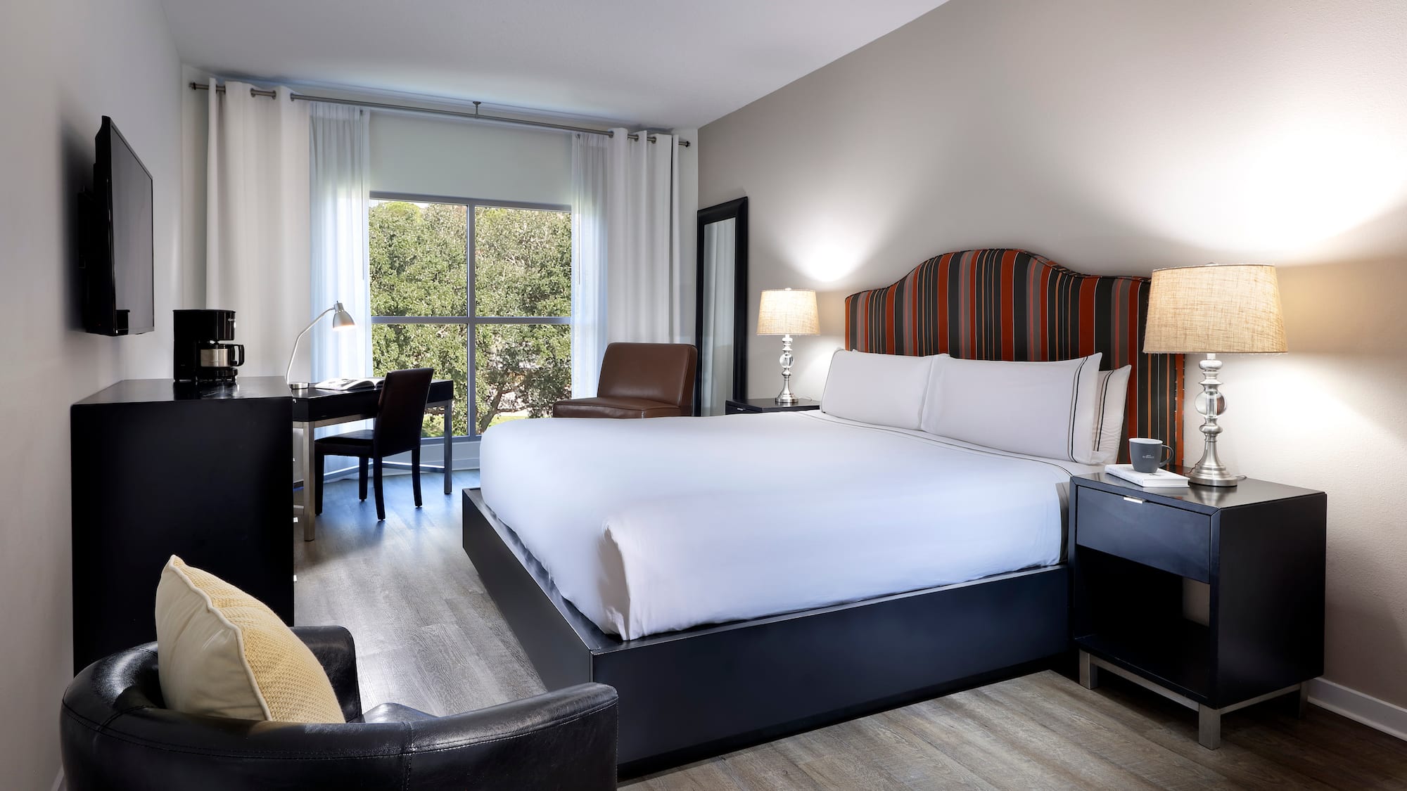 Modern hotel room with king bed, striped headboard, work desk, and window view.