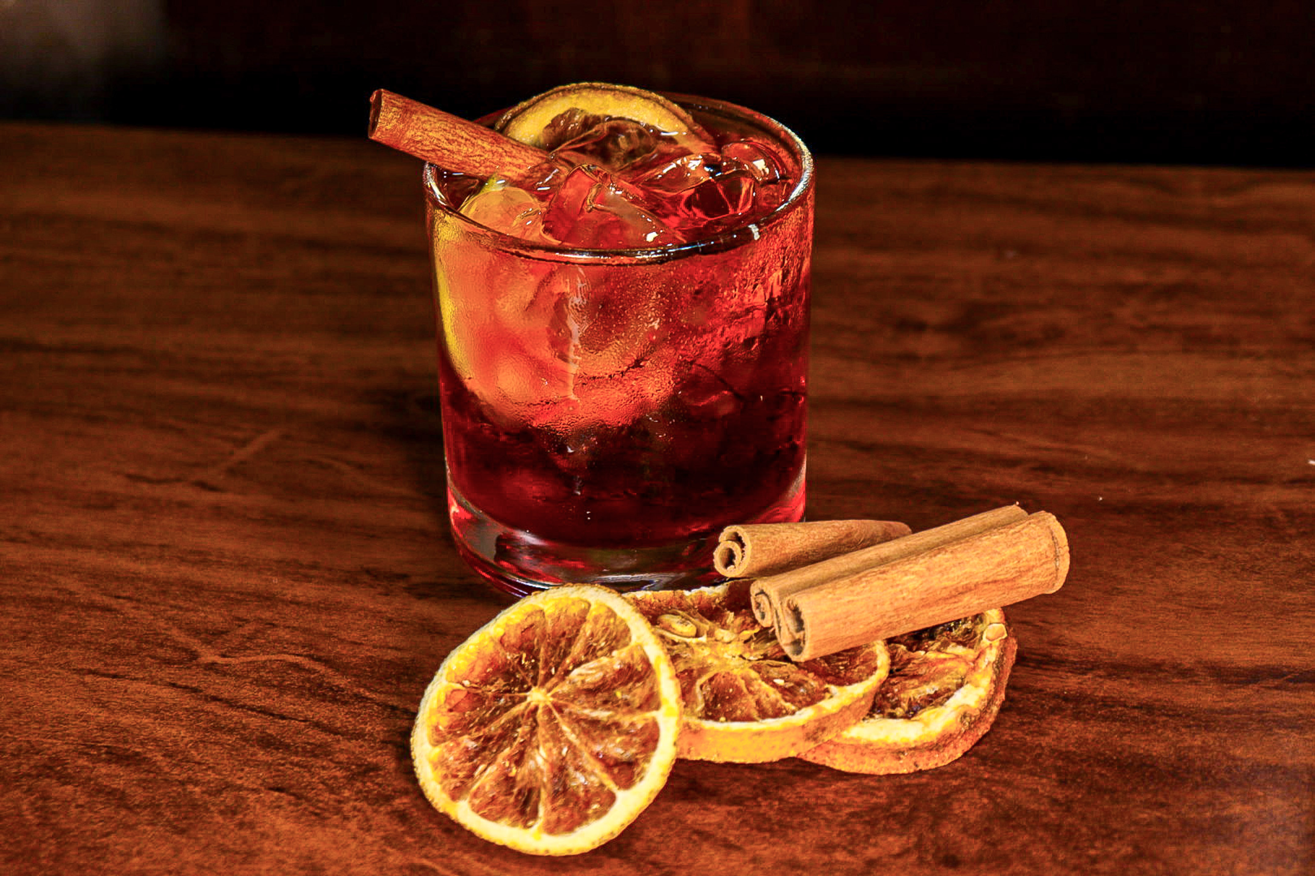 Dark red cocktail with ice, dried orange slice, and cinnamon stick on wood.