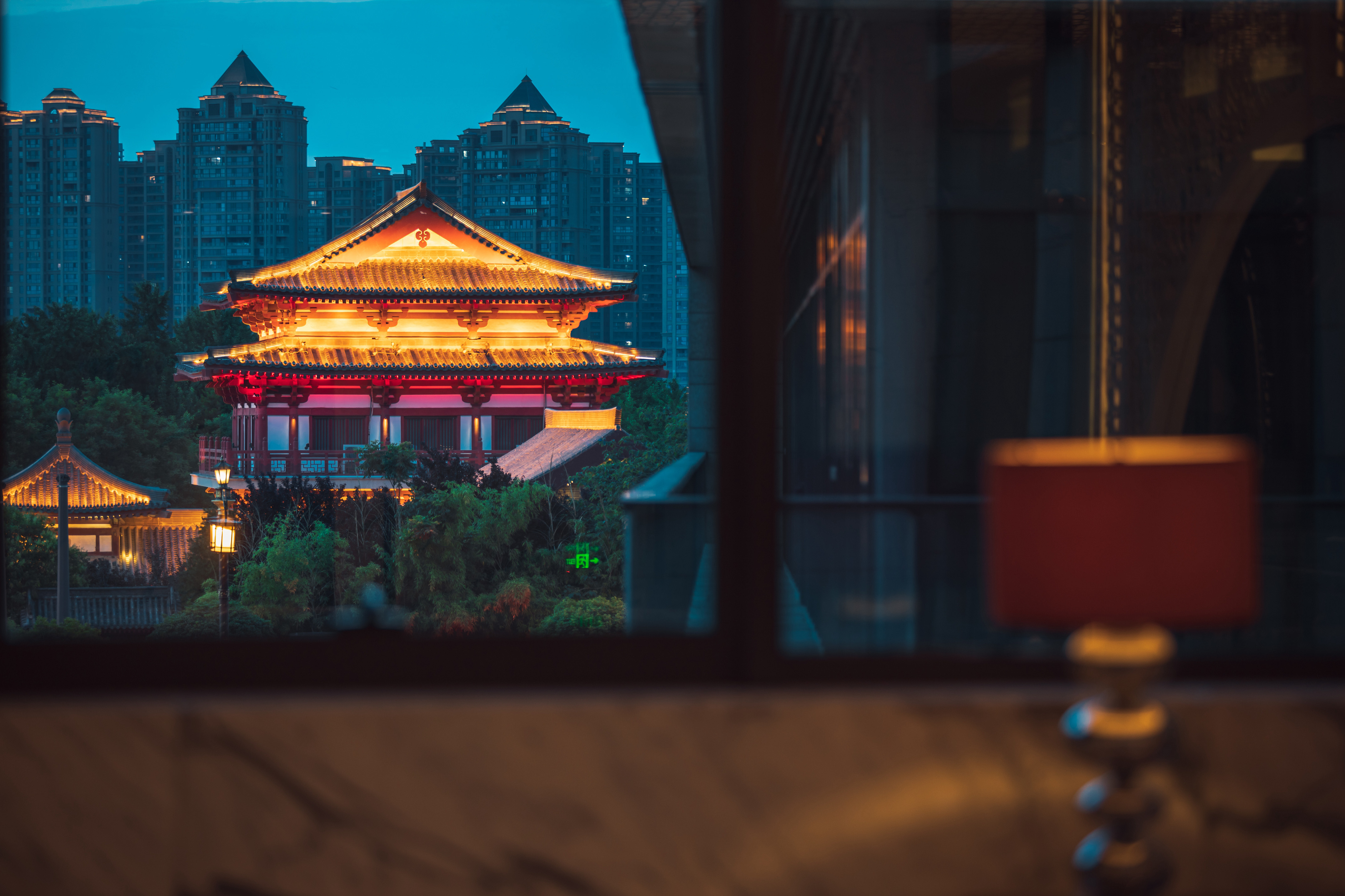Illuminated traditional Chinese pavilion at dusk with city skyline.