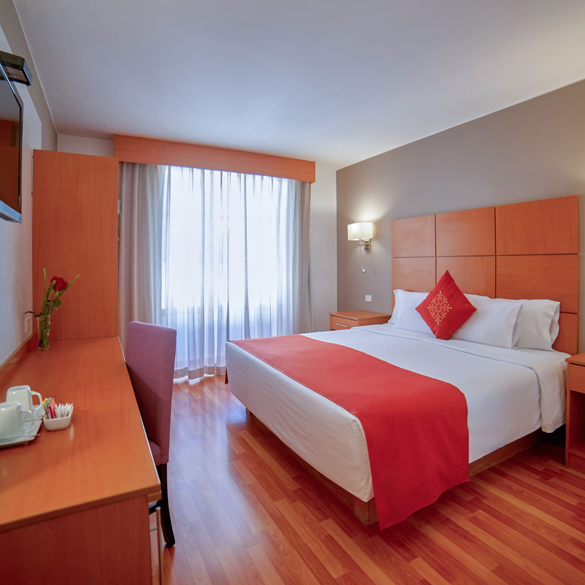 Modern hotel room with bed, red bedding, wooden desk, TV, and bright window.