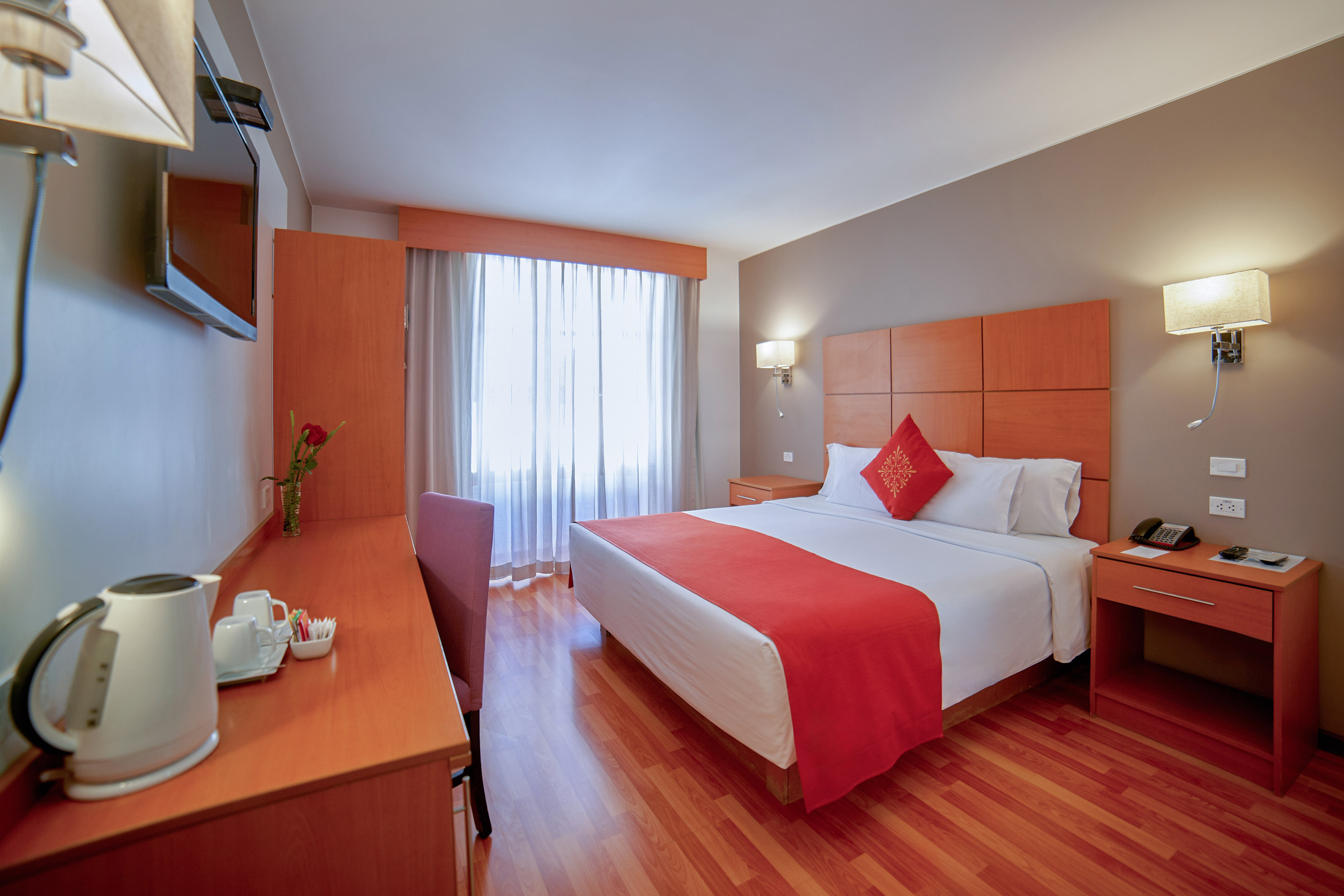 Modern hotel room with bed, red bedding, wooden desk, TV, and bright window.
