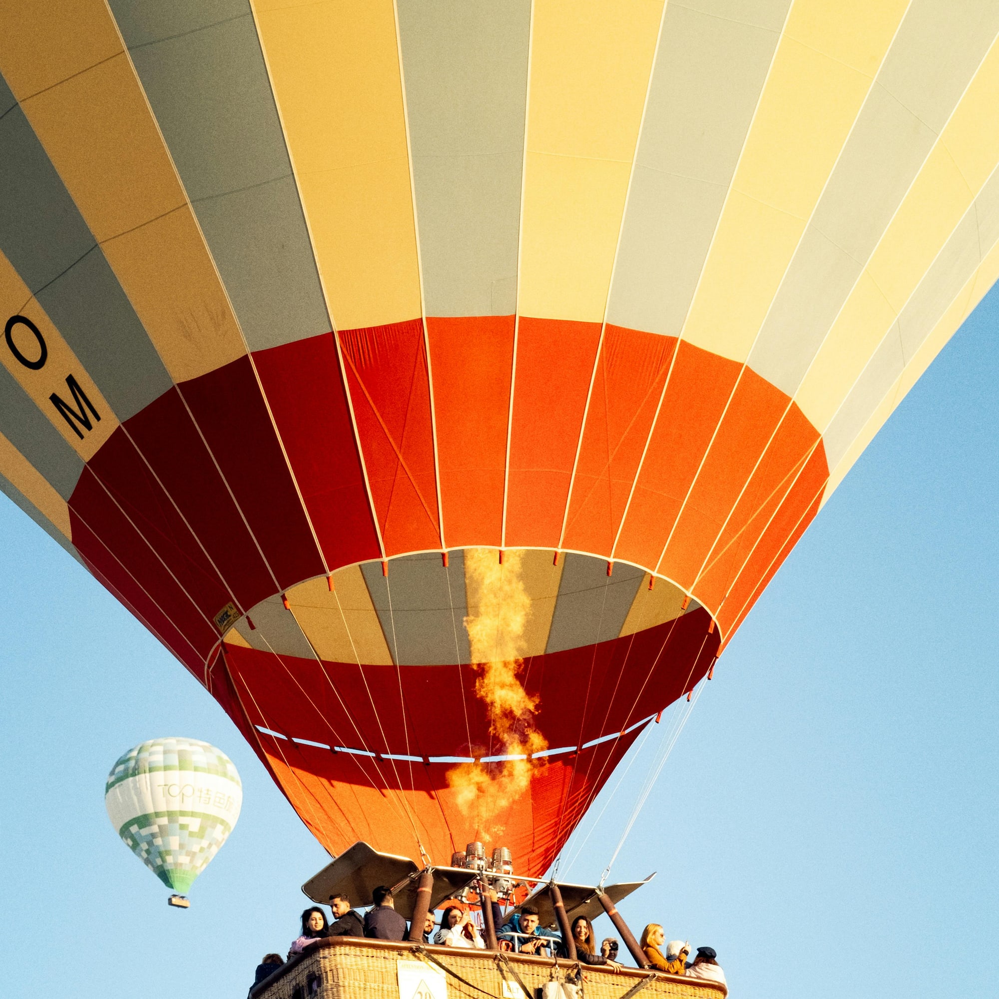 Orange hot air balloon with passengers and flame, smaller balloon in blue sky.