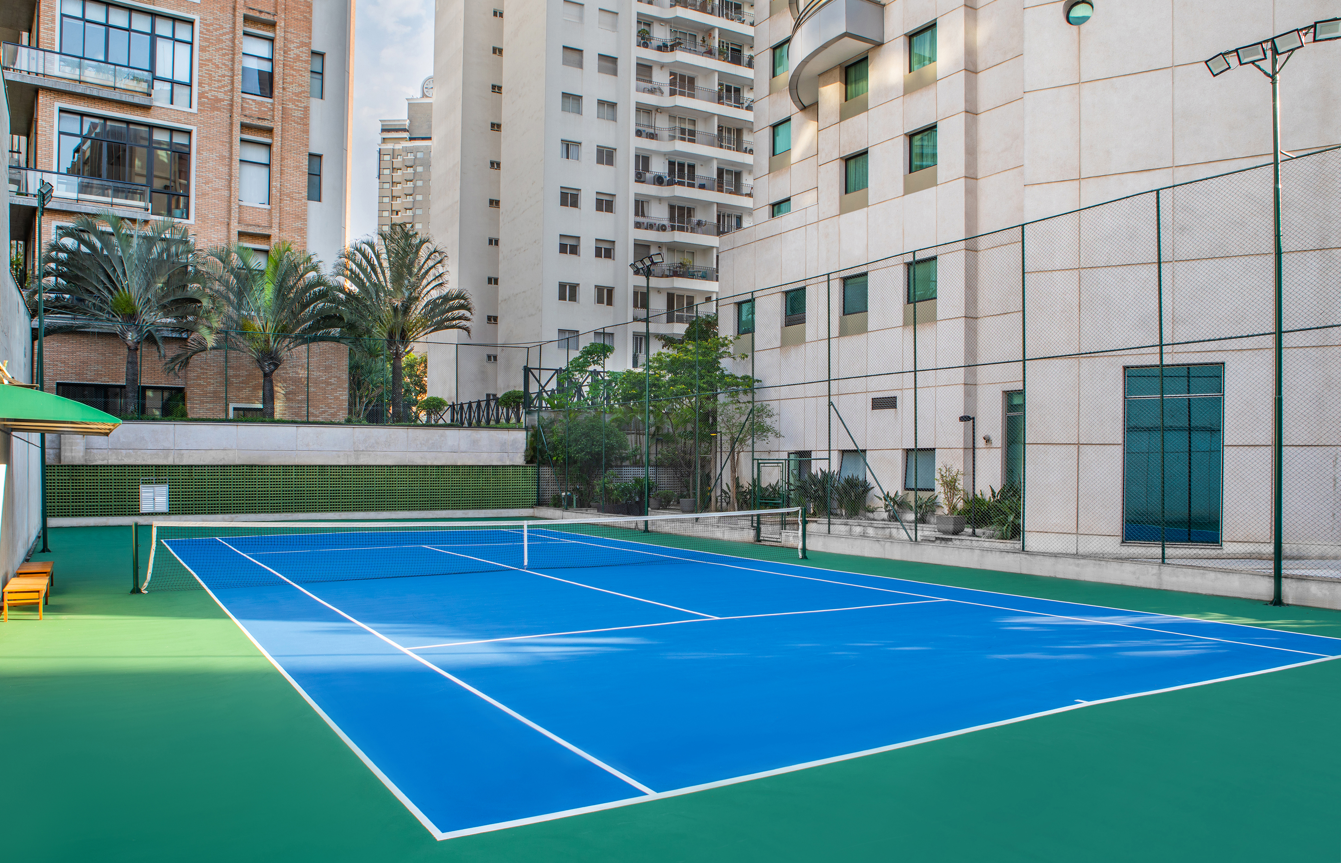 Blue tennis court with green surrounds, modern apartment buildings, and palm trees.