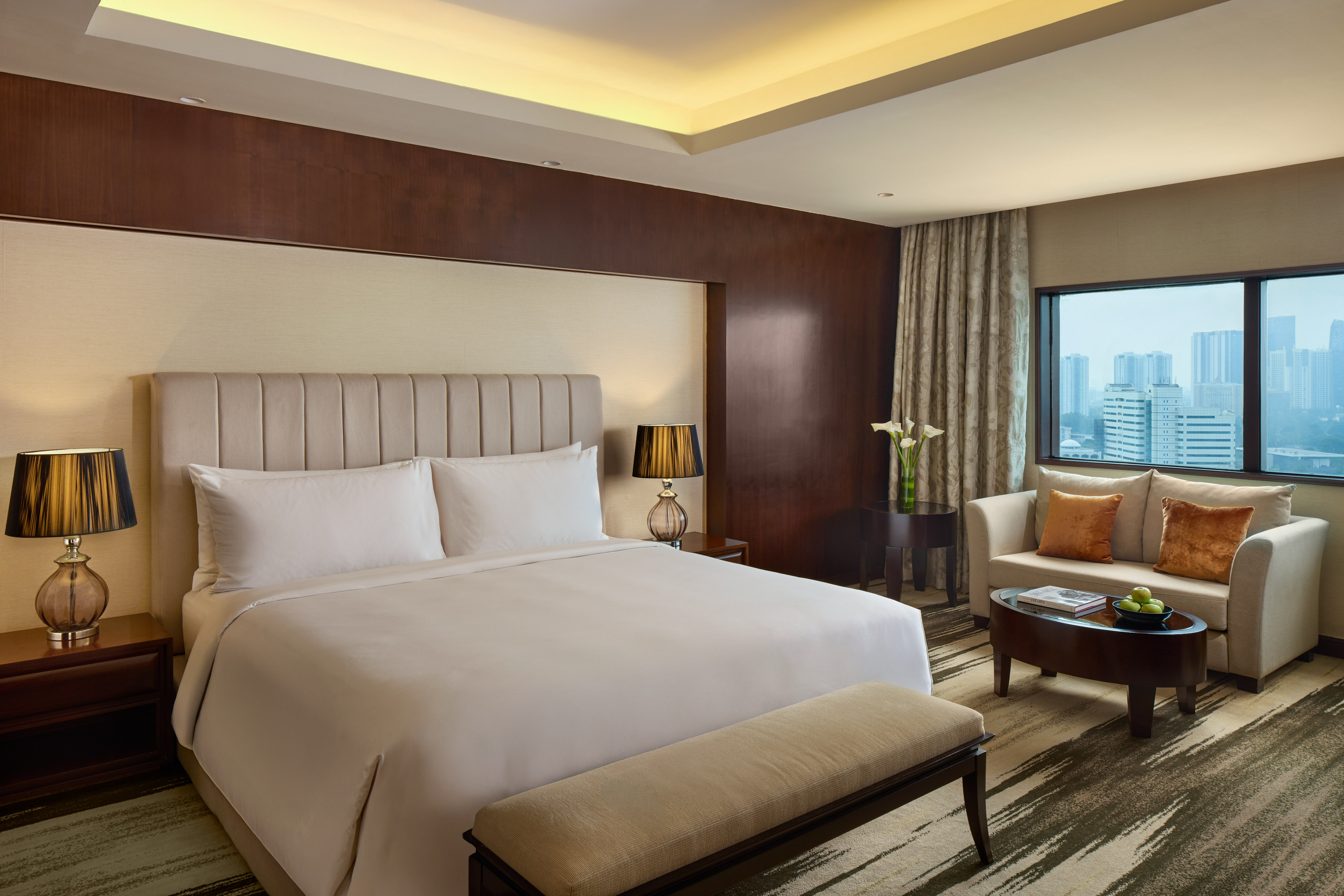 Modern hotel room with king bed, sofa, city view, and elegant wood decor.
