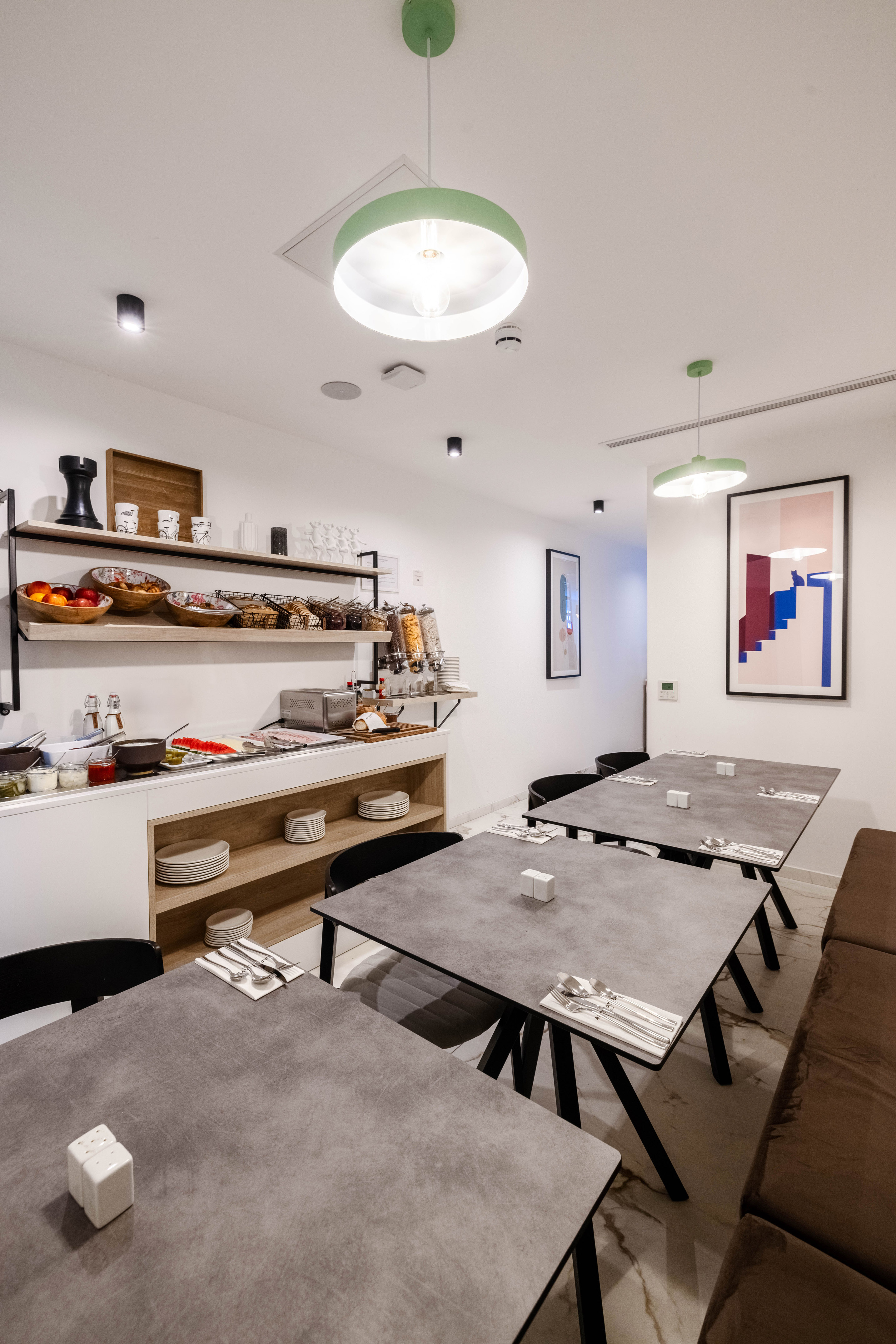 Modern breakfast area with fruit, pastries, cereal shelves, and green pendant lights.