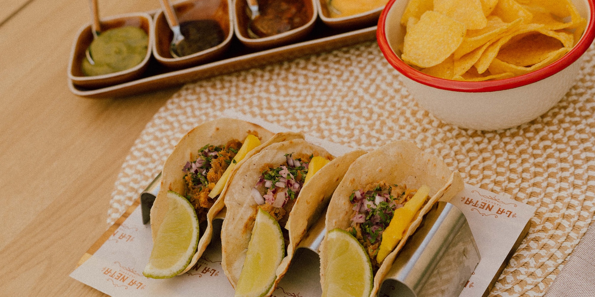 La Neta Taqueria tacos with lime, salsas, and tortilla chips on a wood table.