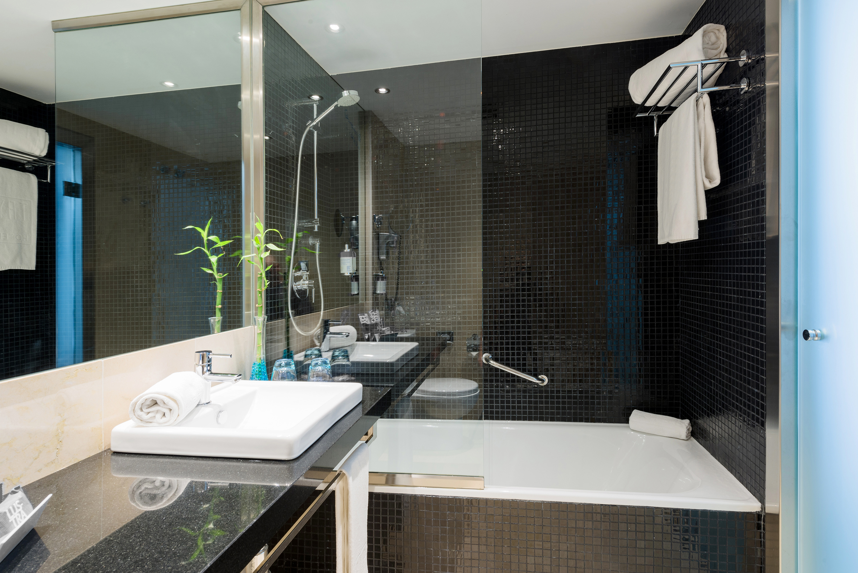Hotel bathroom with black mosaic tiles, white bathtub, glass shower, and vanity.